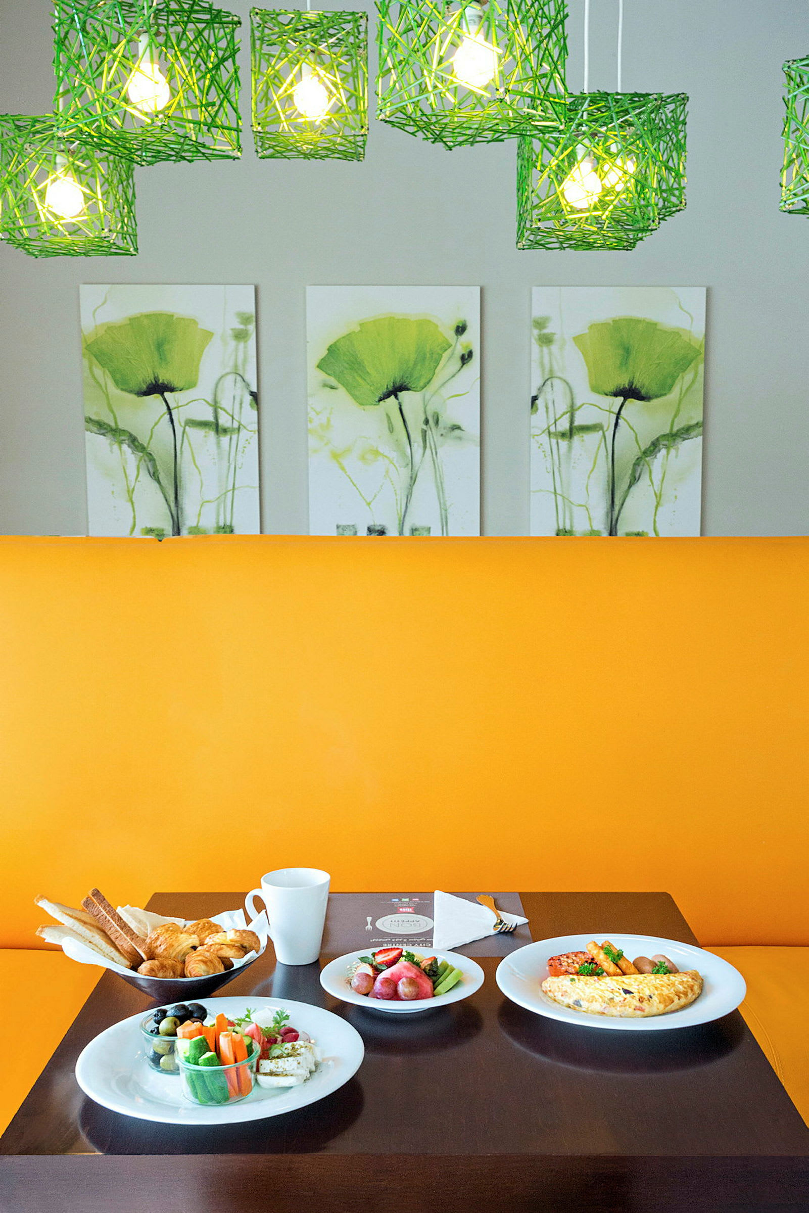 Et bord fylt med frokostmat, inkludert brød, grønnsaker og omelett, foran et gult sete med grønne lamper og blomsterkunst i bakgrunnen. (AI-generert tekst)