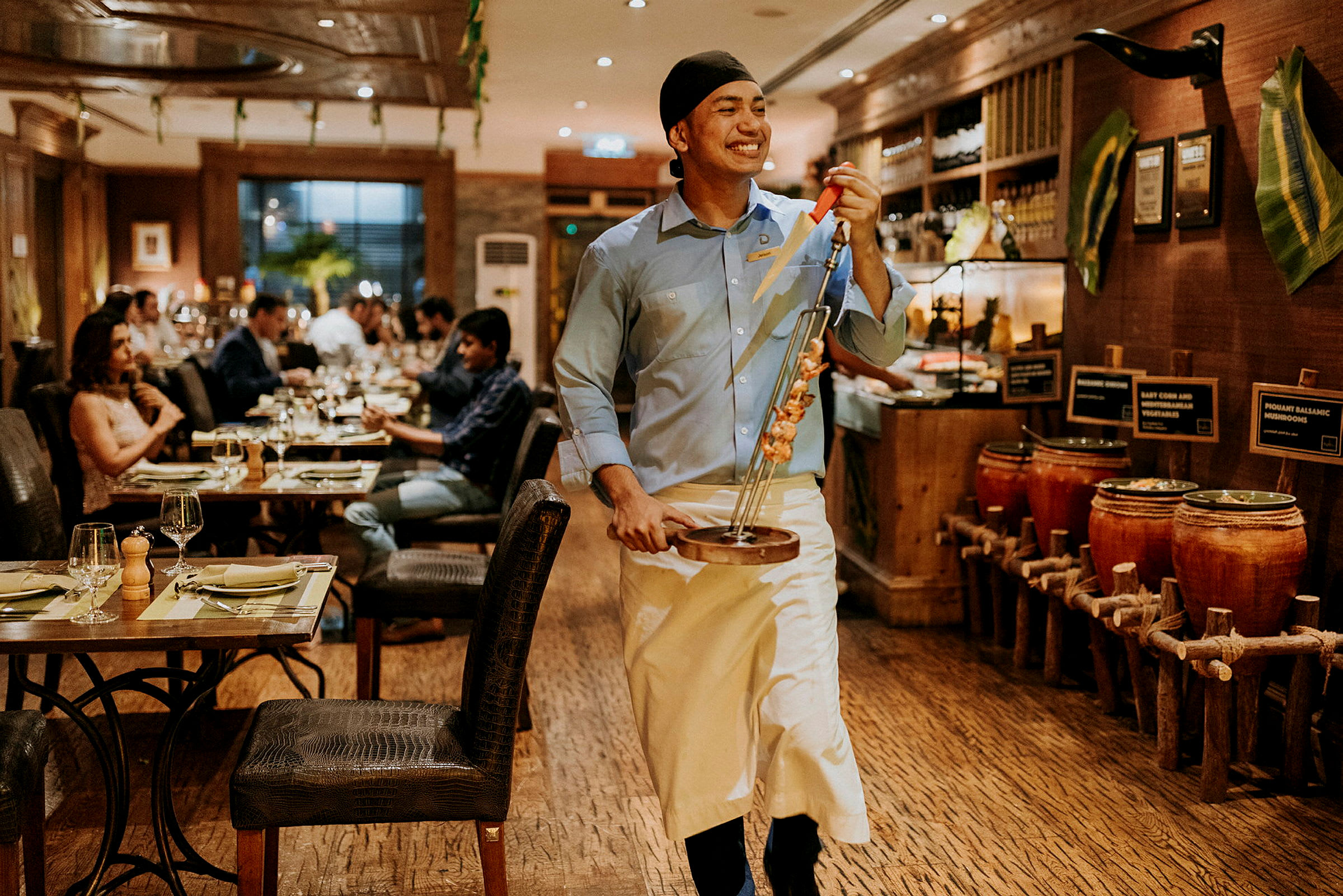 En servitør i en restaurant bærer en tallerken med grillede spyd. Andre gjester sitter ved bord i bakgrunnen. (AI-generert tekst)