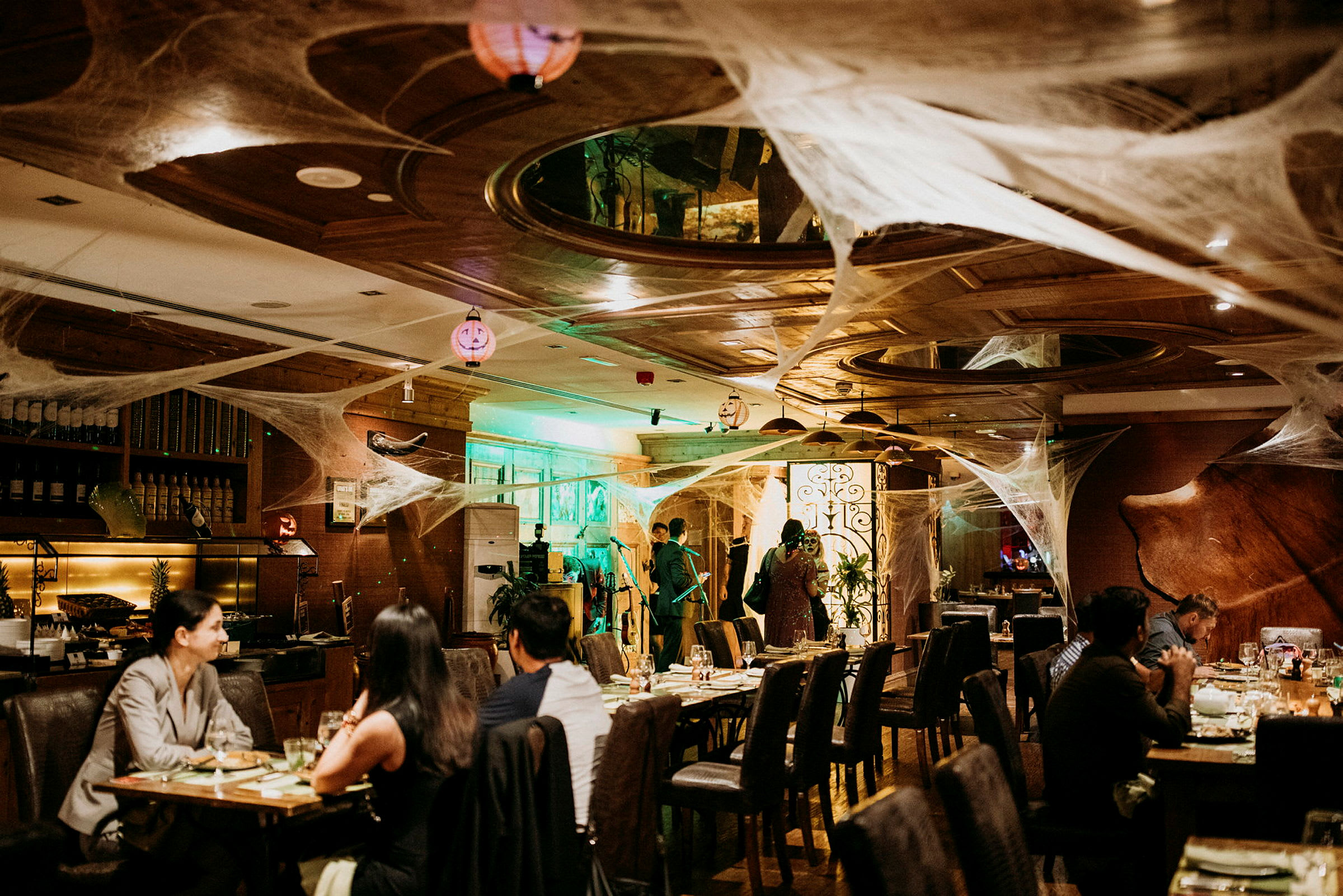 Et restaurantinteriør dekorert med edderkoppnett-lignende pynt og belysning, med gjester ved bord og personale i bakgrunnen. (AI-generert tekst)