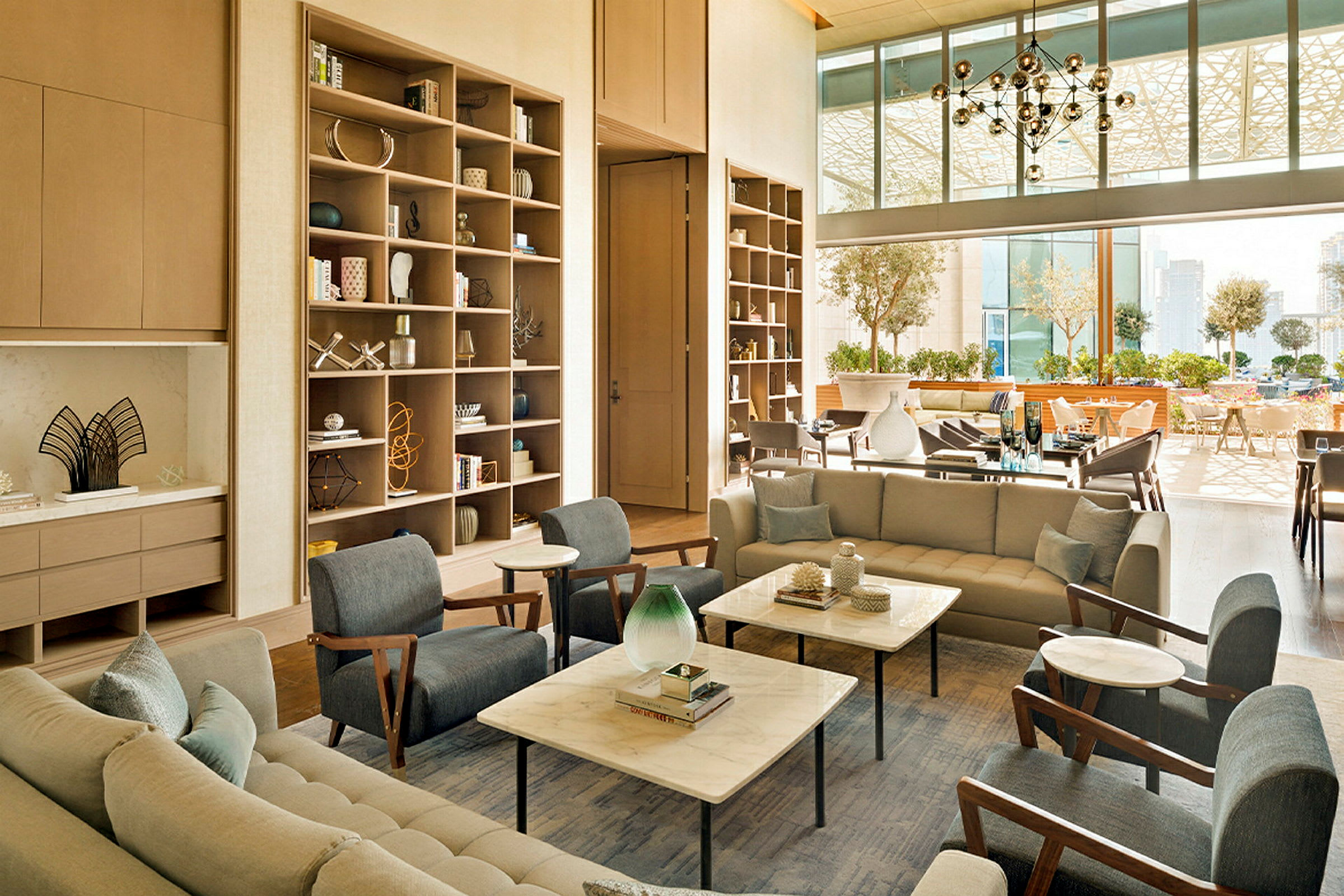 Et lyst og moderne loungemiljø med komfortable sofaer, lenestoler, marmorbord og et åpent hylle-system. I bakgrunnen sees store vinduer og en terrasse med stoler og bord. (AI-generert tekst)