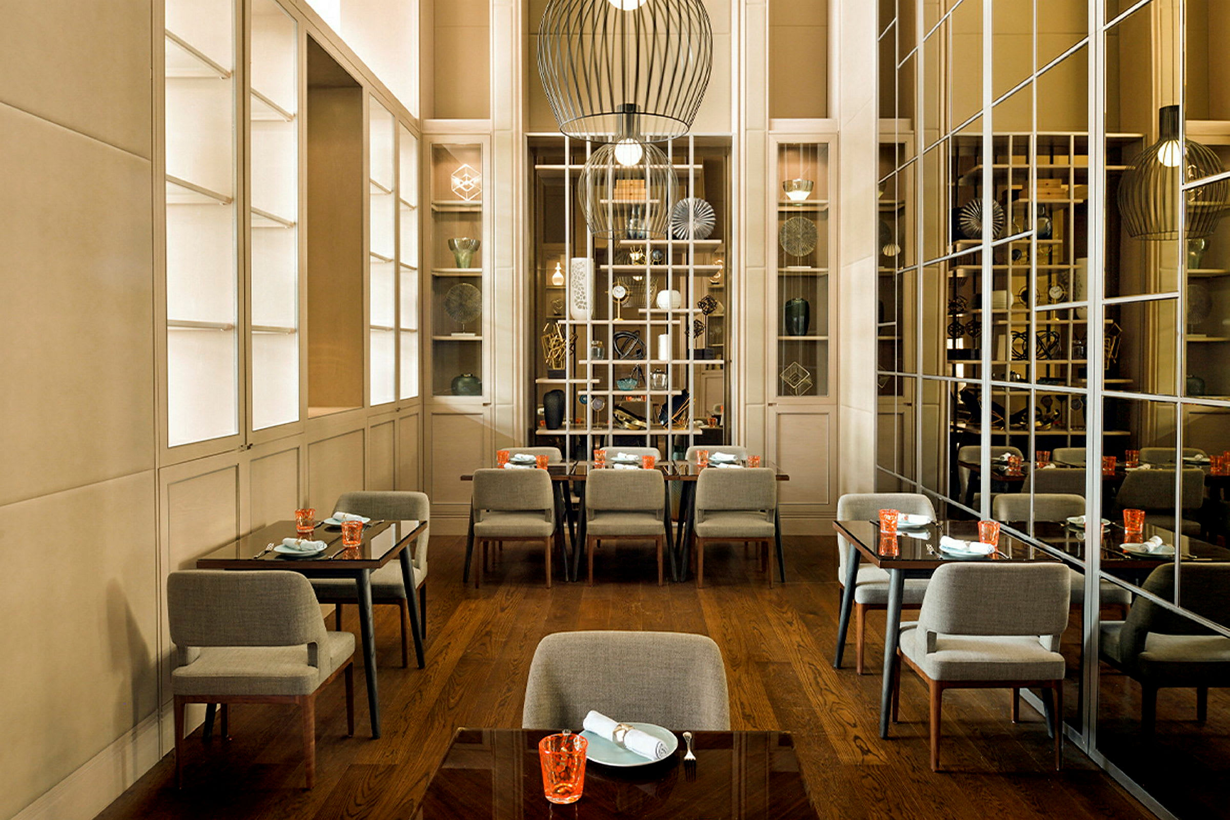 Et elegant innredet restaurantområde med flere bord og stoler, speilvegger, hyller med dekorative gjenstander og moderne hengende lamper. (AI-generert tekst)