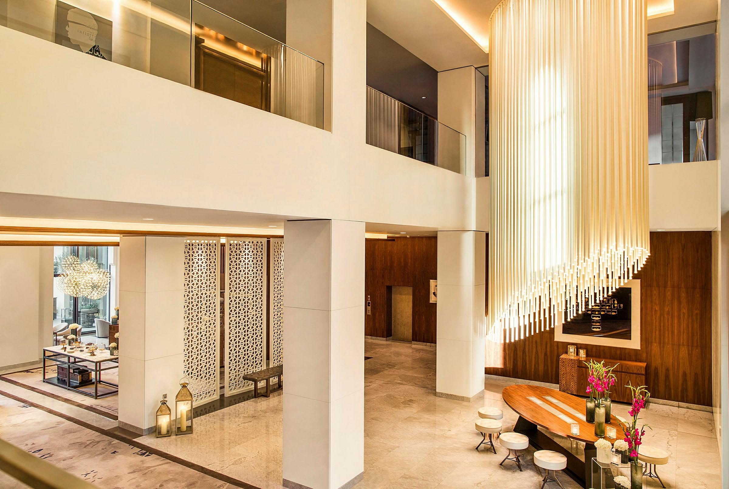 En elegant hotellobby med høy takhøyde, moderne interiør og en lysekrone laget av vertikale rør. Det finnes sitteplasser rundt et trebord med blomster. (AI-generert tekst)