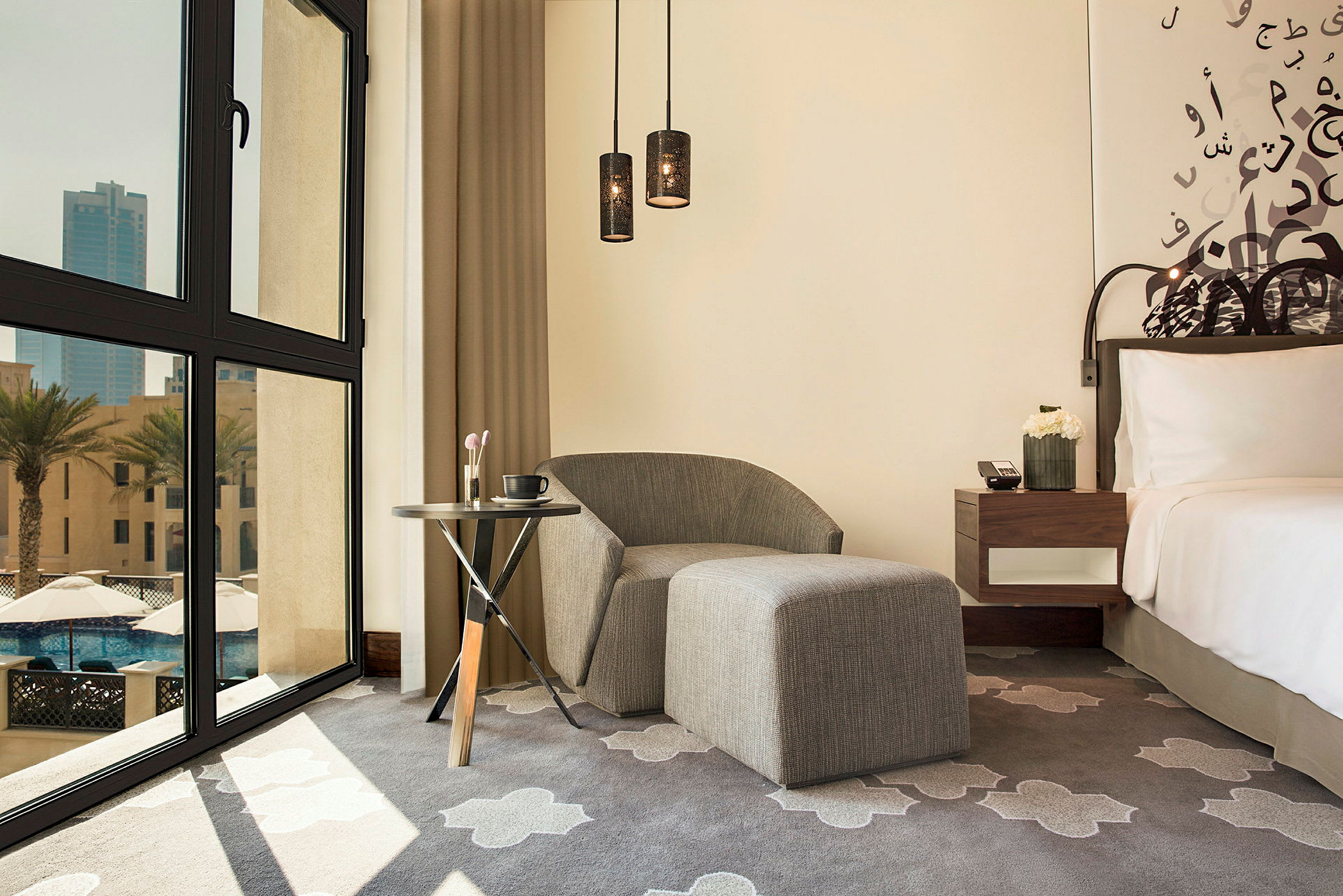Et hotellrom med moderne innredning. Bildet viser en grå stol med fotpall, et lite bord med en kaffekopp og sminkekoster, samt en seng med hvite sengeklær. Et stort vindu med utsikt over palmetrær og bygninger er også synlig. (AI-generert tekst)