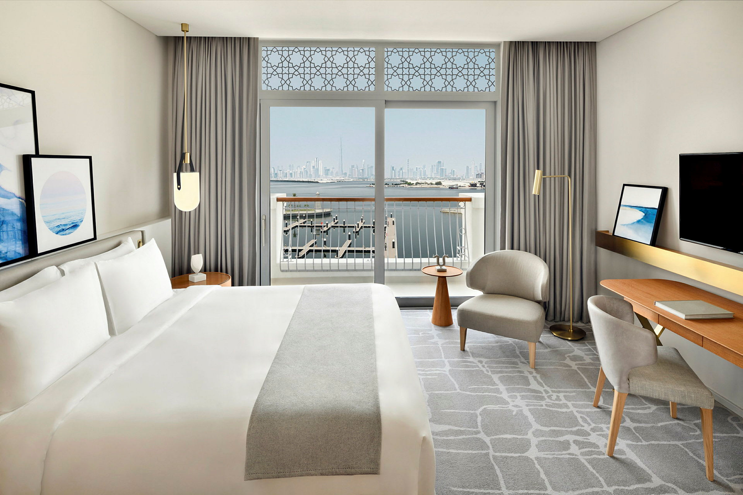 Hotellrom med en stor seng, skrivebord, sitteplasser og en balkong med utsikt over vann og en bysilhuett. (AI-generert tekst)
