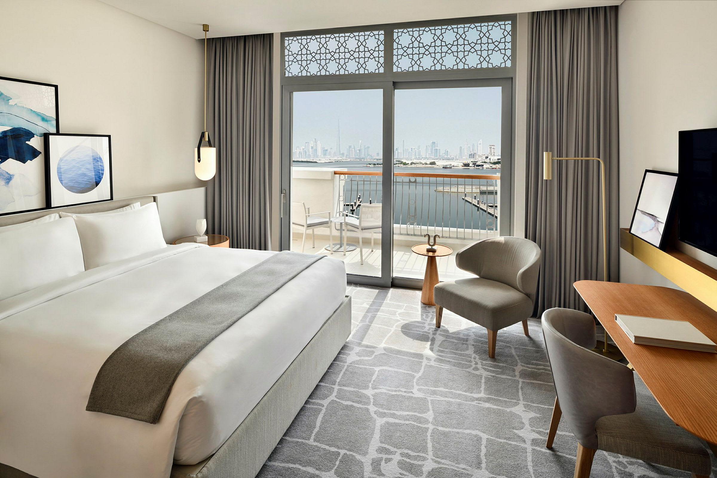 Et moderne innredet hotellrom med en stor dobbeltseng og vinduer som åpner opp til en balkong med utsikt over en bysilhuett og vann. (AI-generert tekst)