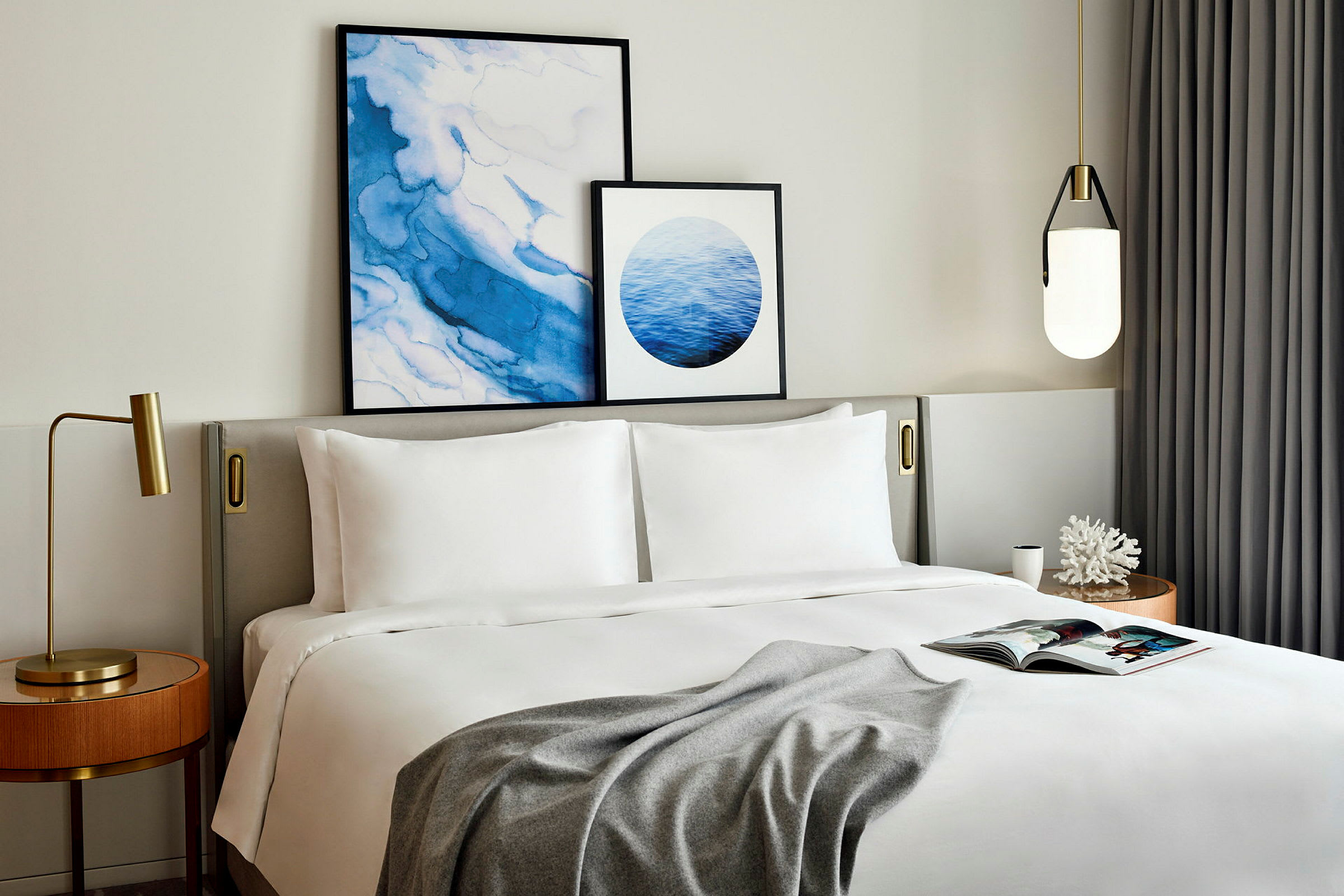 Et elegant innredet soverom med en stor seng, dekorert med hvitt sengetøy og et grått teppe. På sengegavlen henger to abstrakte kunstverk i blå og hvite nyanser. På et nattbord står en gullfarget lampe, og på det andre står en pyntegjenstand i form av en hvit korall. (AI-generert tekst)