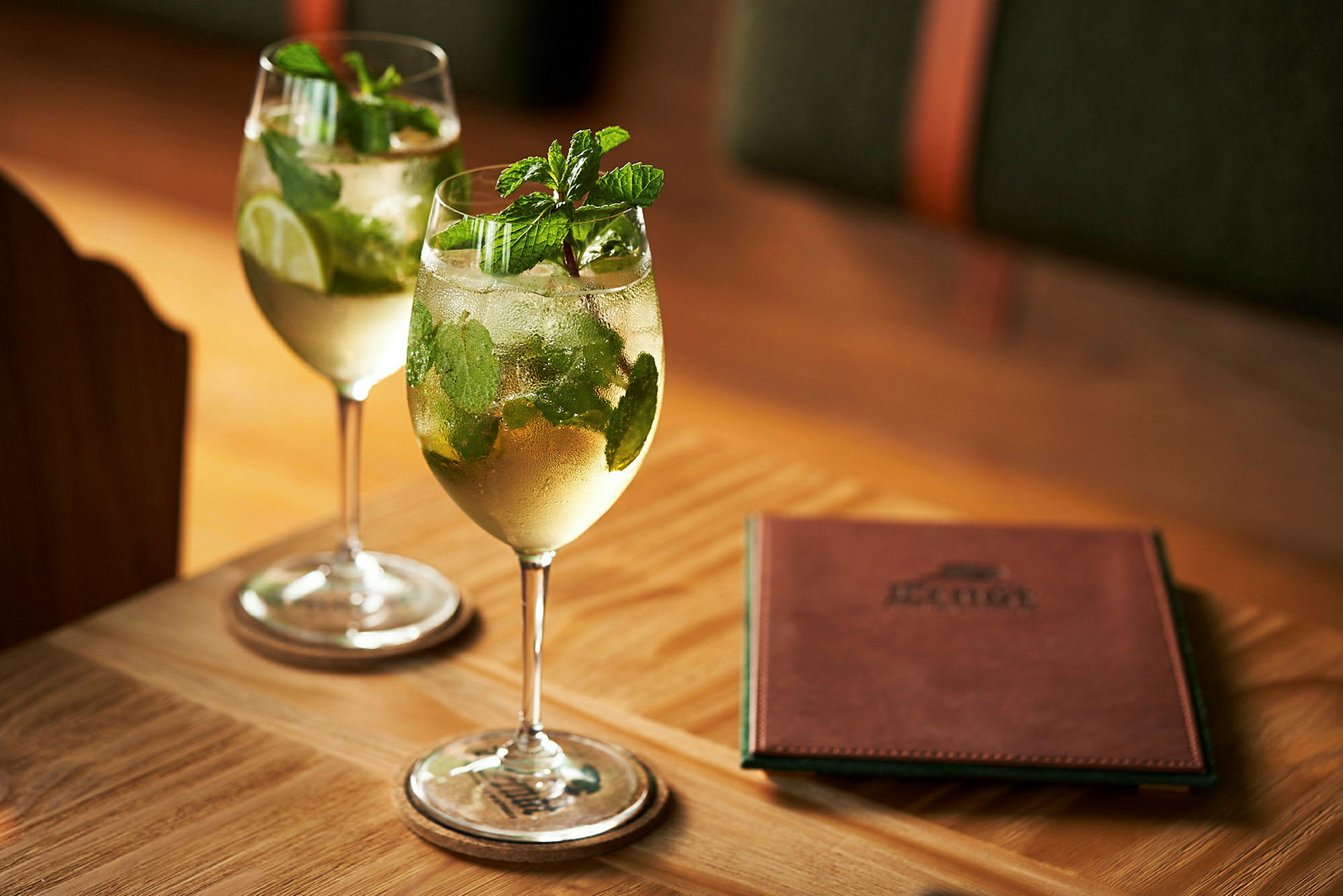 To glass med drikker som inneholder mynte og lime står på et bord ved siden av en menyhefte. (AI-generert tekst)