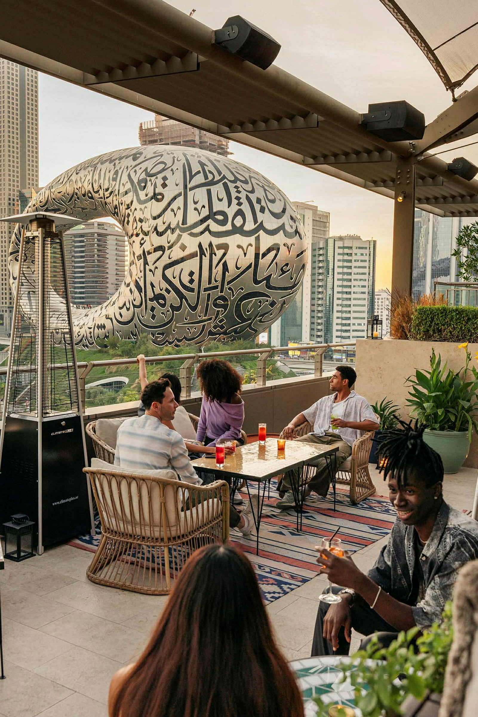 Personer som sitter ved et bord på en takterrasse med Museum of the Future i bakgrunnen. (AI-generert tekst)