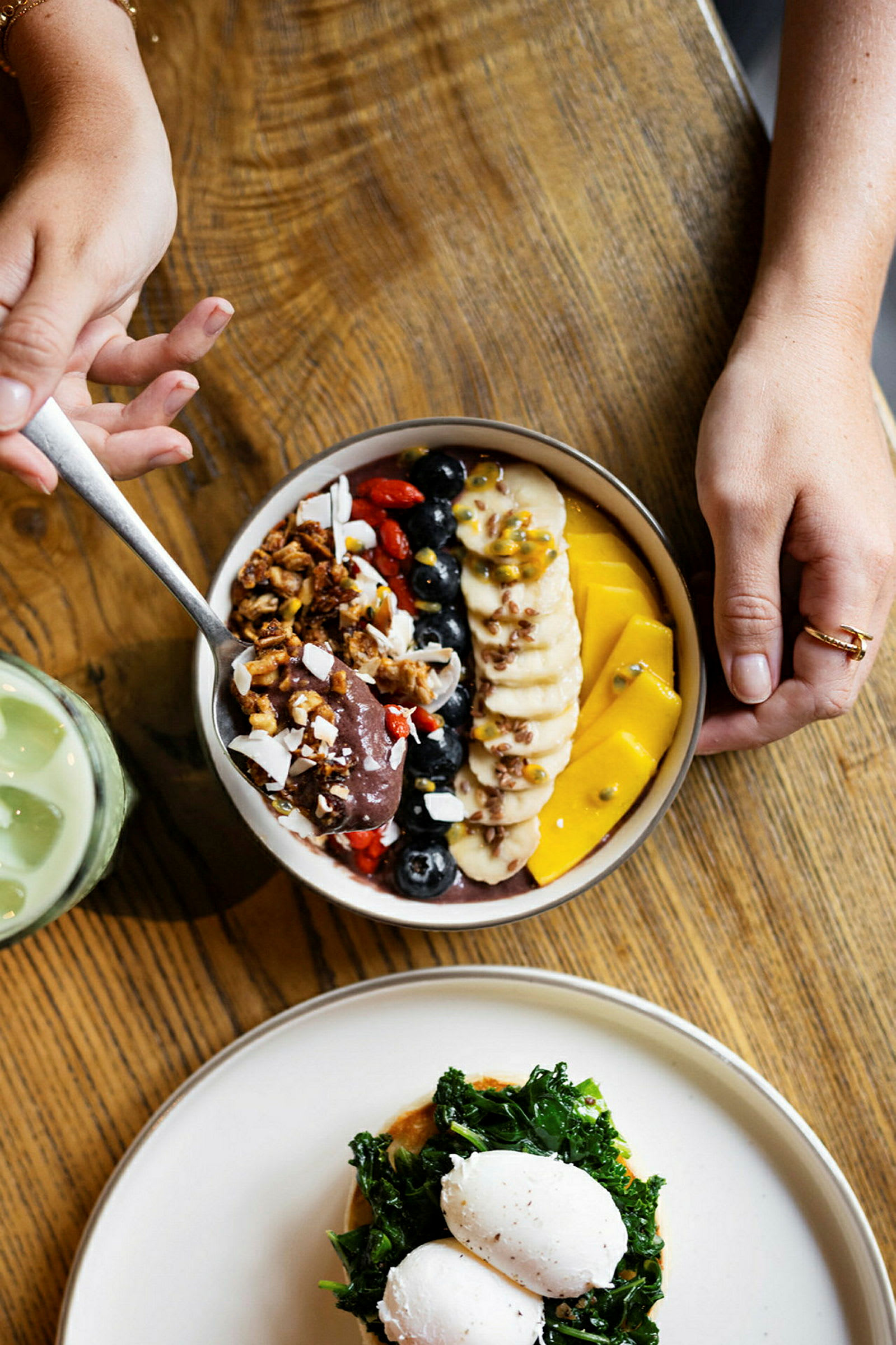 En tallerken med en fargerik acai-bolle og en annen med posjerte egg og grønnsaker på et trebord. (AI-generert tekst)