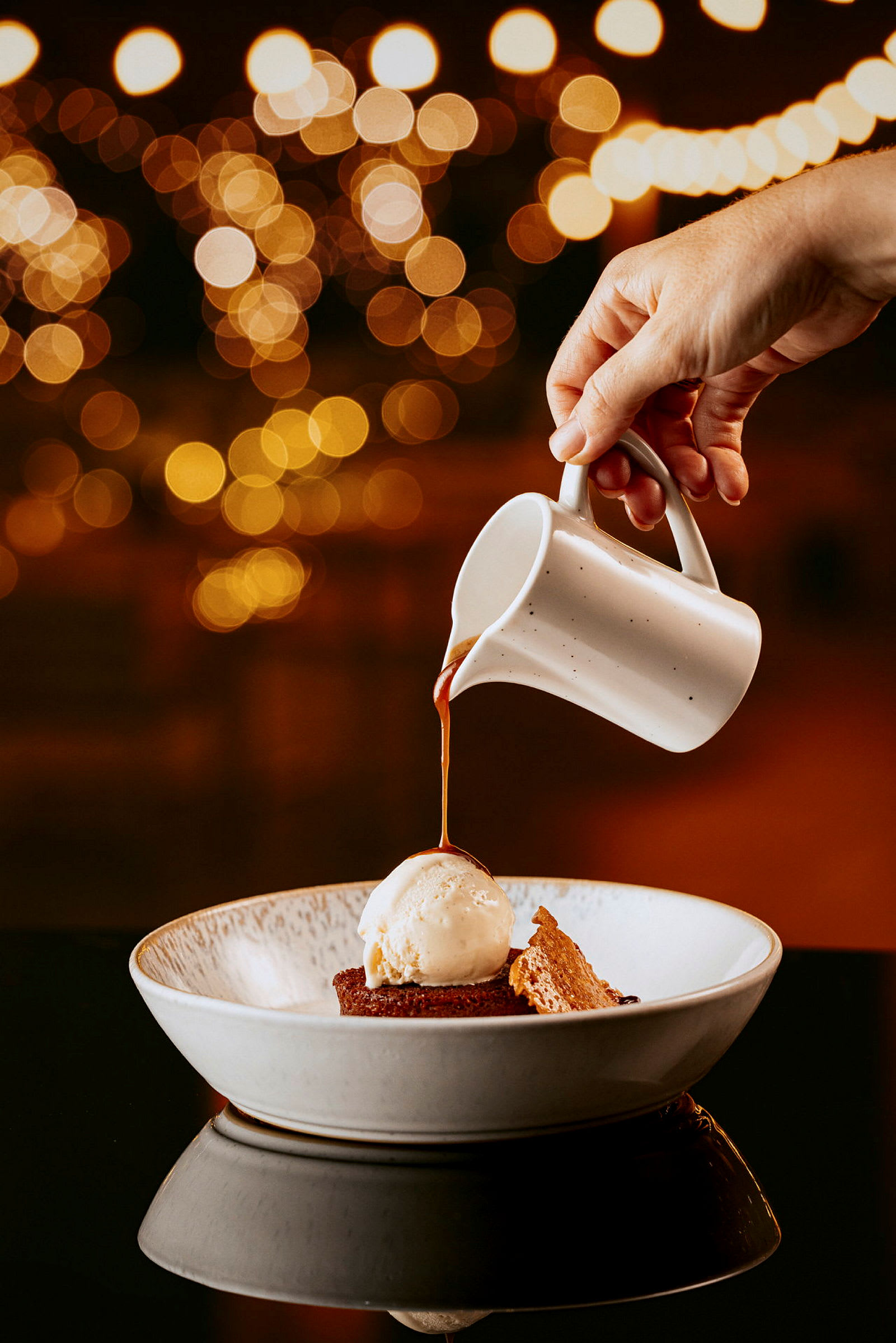 En hånd heller saus fra en liten kanne over en dessert med iskrem og kake i en skål, med uskarpt lys i bakgrunnen. (AI-generert tekst)