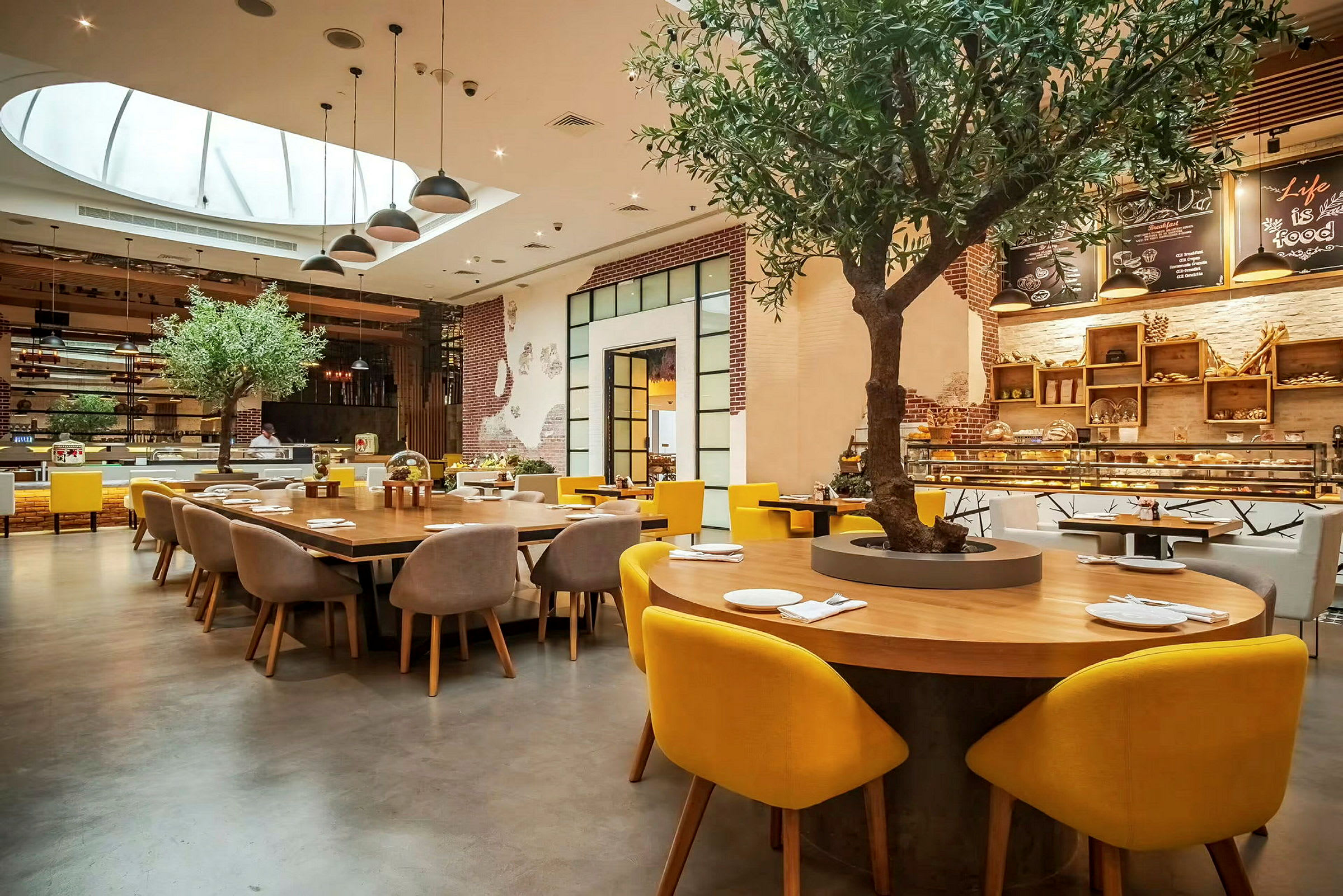 Interiør av en restaurant med et stort rundt bord omgitt av stoler med gule ryggstøtter. Et tre er plantet midt i bordet, og bakgrunnen viser en serveringsdisk og dekorasjoner. (AI-generert tekst)