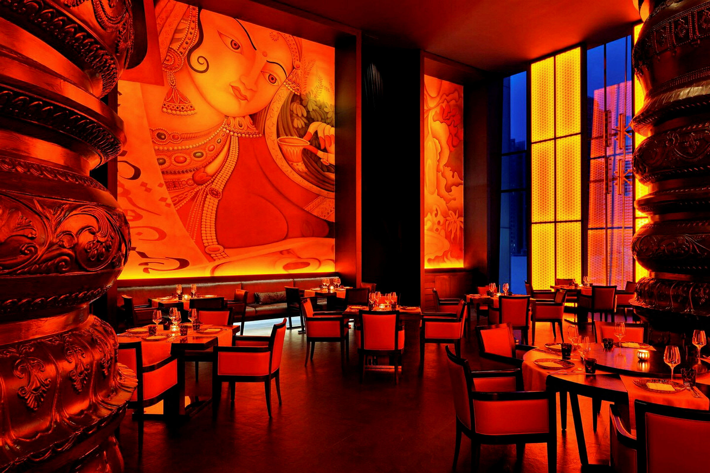 En restaurant med røde og oransje fargetemaer, høye vegger med kunst og store vinduer. Møblene består av bord og stoler. (AI-generert tekst)