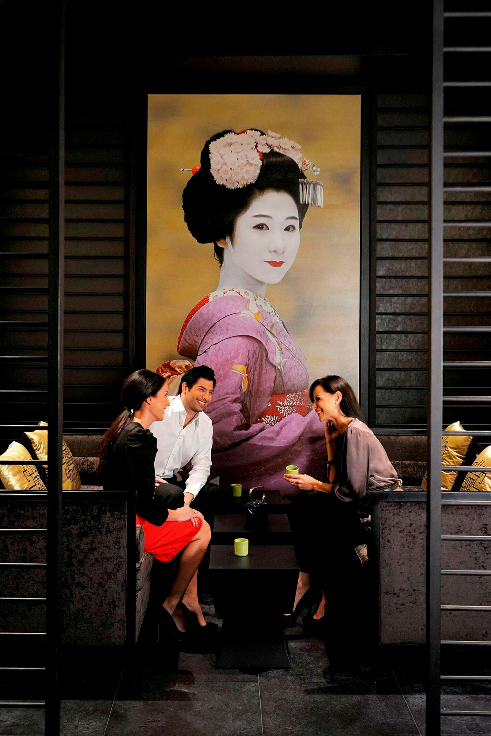 Tre personer sitter ved et bord i en elegant setting med et stort bilde av en geisha i bakgrunnen. (AI-generert tekst)