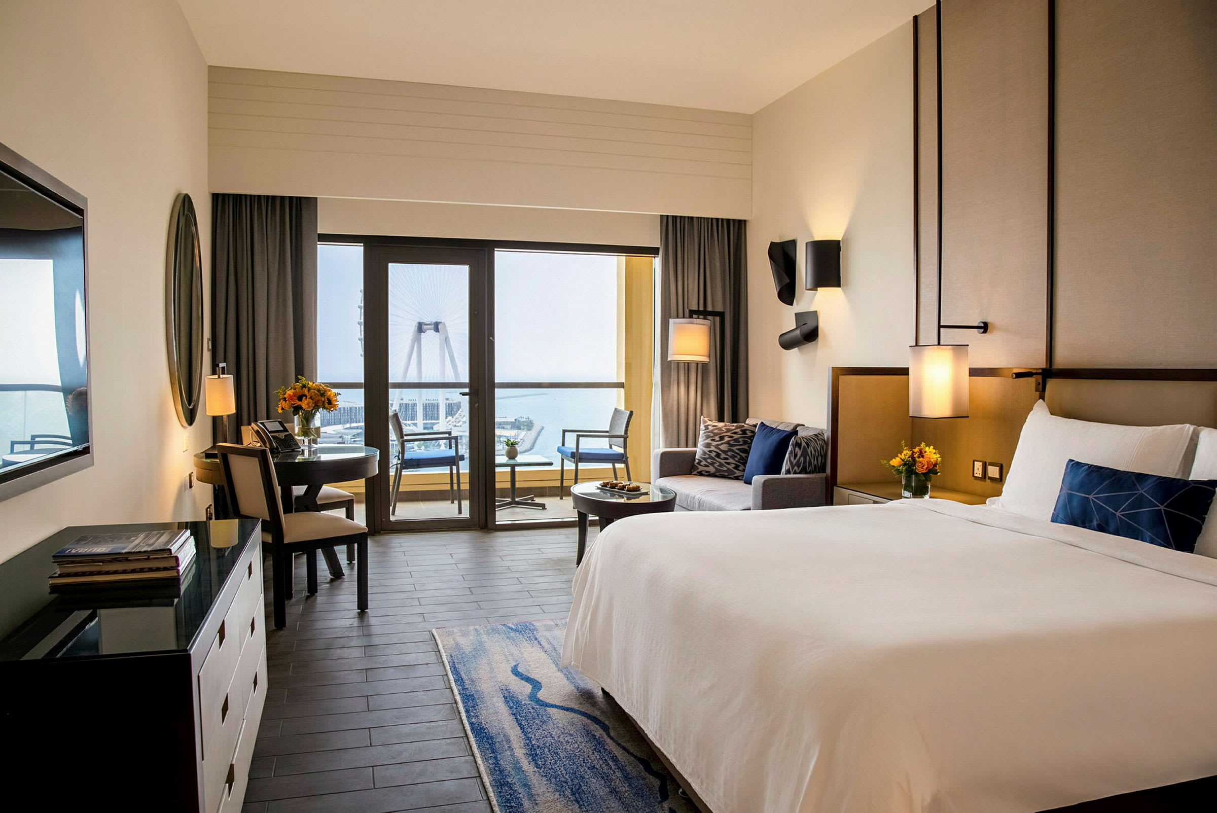 Et hotellrom med en stor seng, sittegruppe og en spiseplass. Store vinduer leder ut til en balkong med utsikt over havet og et stort pariserhjul. (AI-generert tekst)