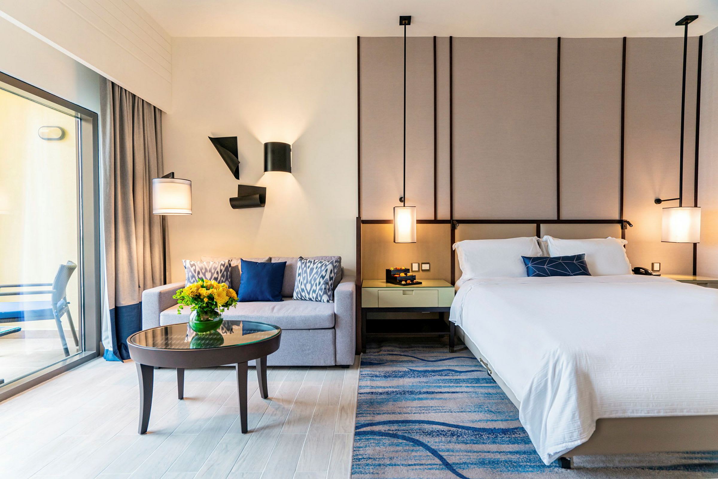 Et moderne hotellrom med en seng, en sofa, et lite sofabord med en vase med blomster og en balkong som er synlig gjennom en glassdør. (AI-generert tekst)