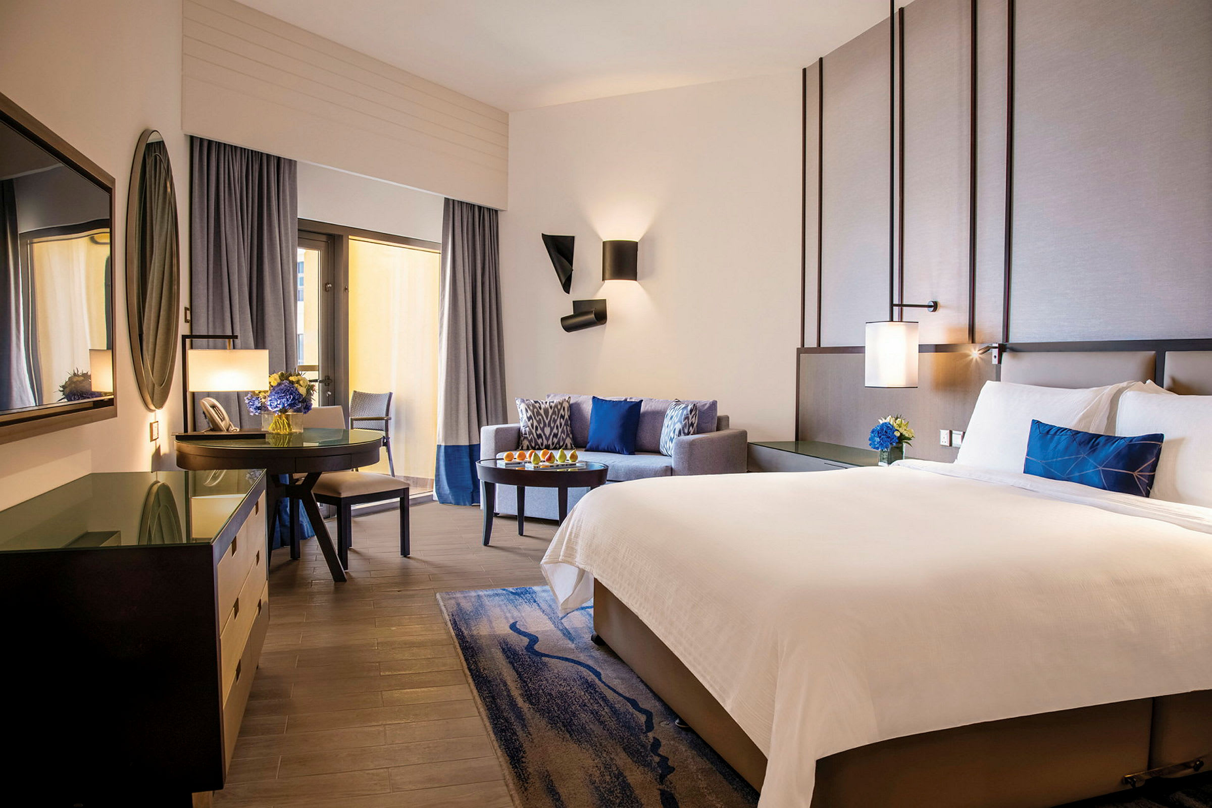 Hotellrom med en stor dobbeltseng, sittegruppe, skrivebord og en TV. Rommet er elegant innredet med nøytrale farger og blå detaljer. (AI-generert tekst)