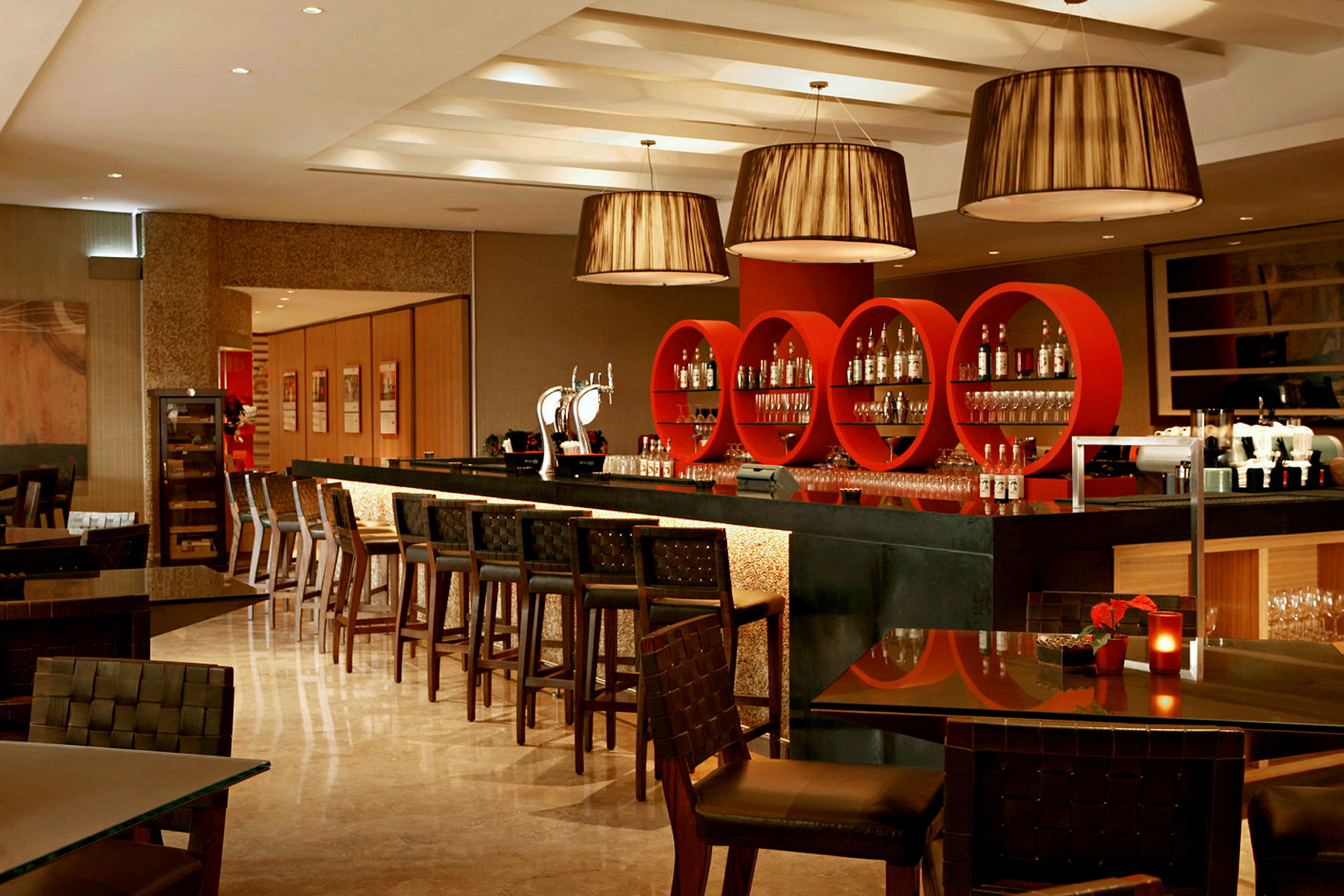 Bilde av en moderne bar med røde, sirkelrunde hyller og belysning. Bartenderdisk og barstoler, samt bord med stoler i forgrunnen. (AI-generert tekst)