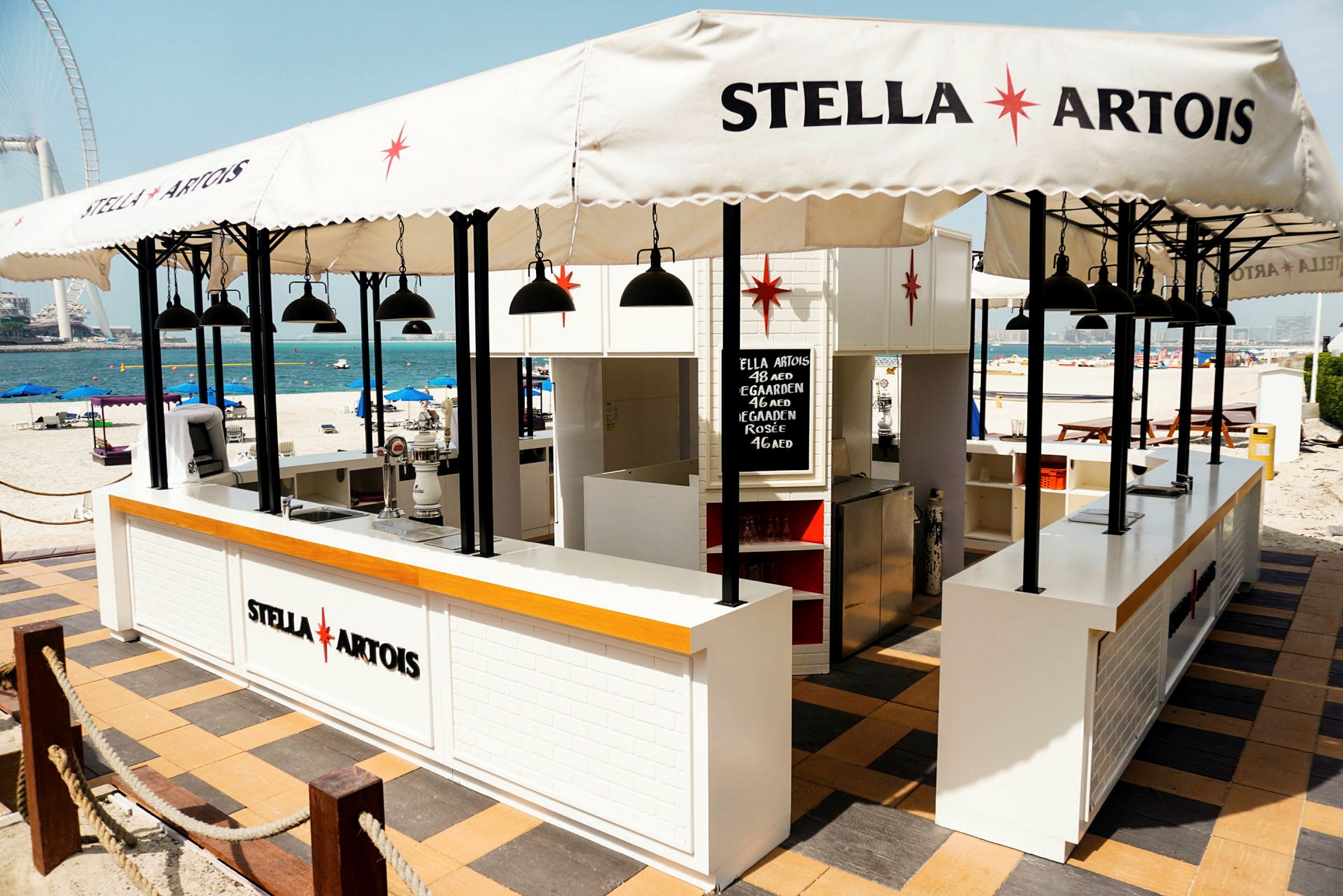 En strandbar med Stella Artois-logo, parasoller og bord. (AI-generert tekst)