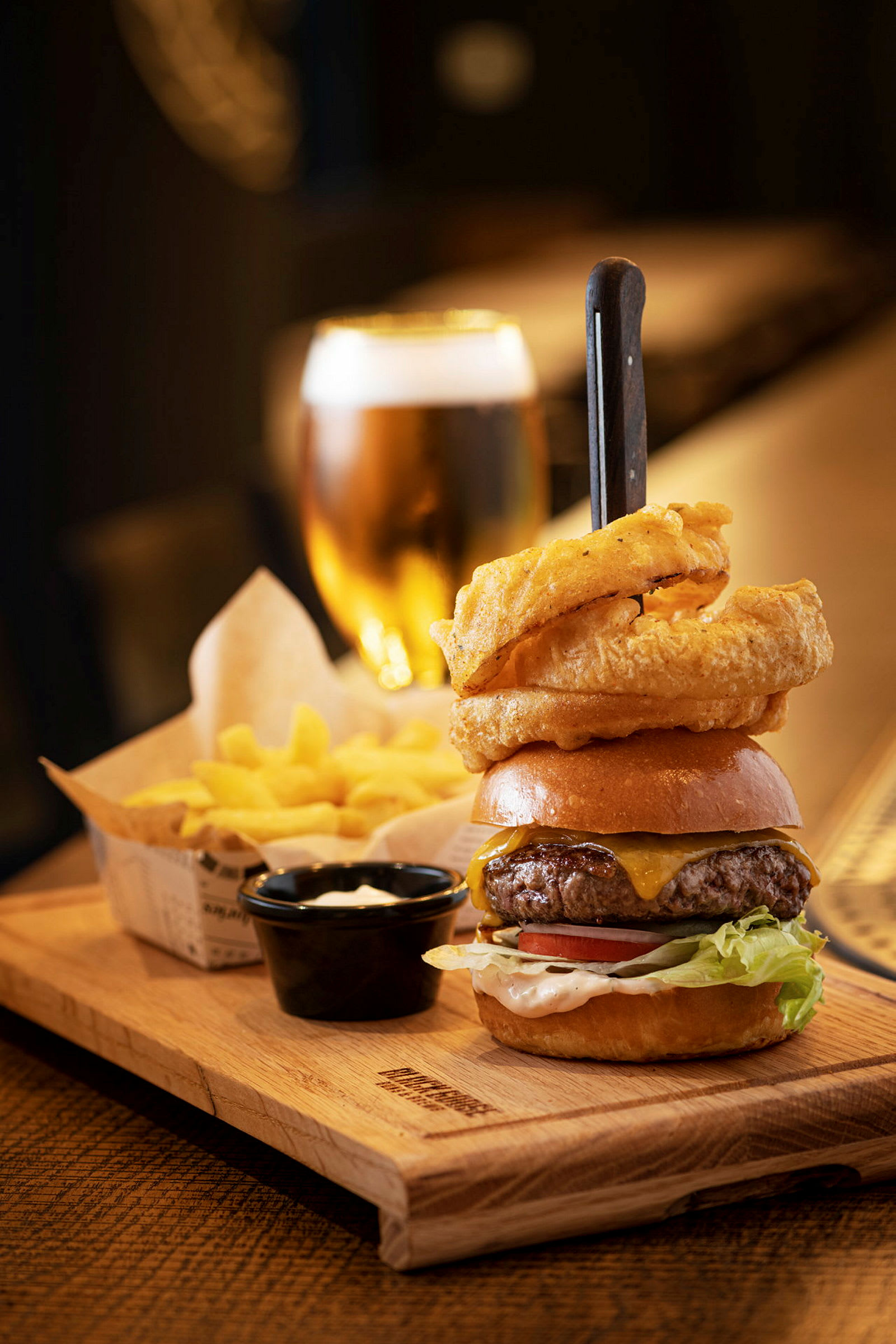 En tallerken med en hamburger, løkringer, pommes frites og en dipp. En øl i glass vises i bakgrunnen. (AI-generert tekst)
