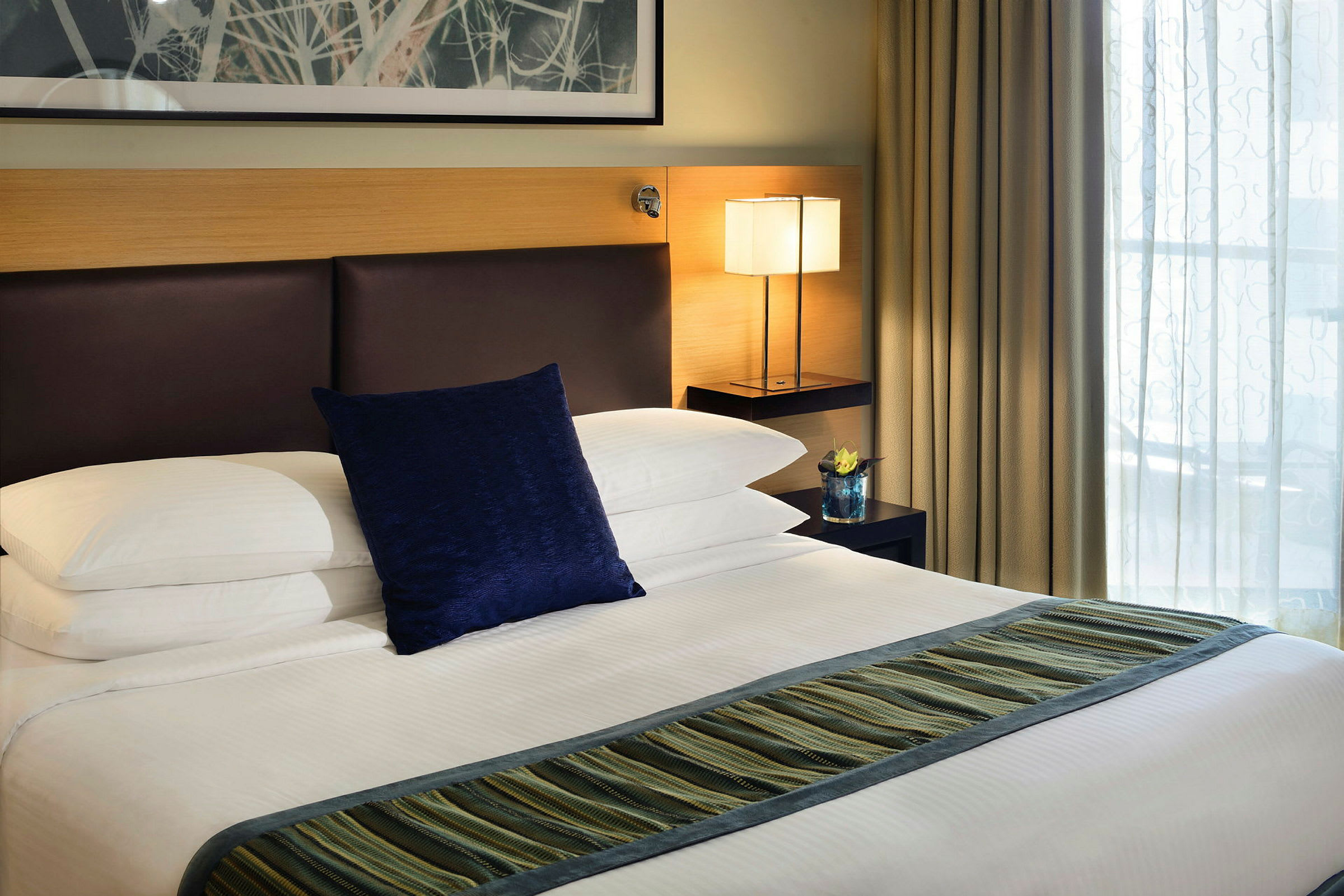En hotellseng med hvitt sengetøy, en blå pute og en stripete løper. Et nattbord med en lampe og en dekorasjon er plassert ved siden av. (AI-generert tekst)
