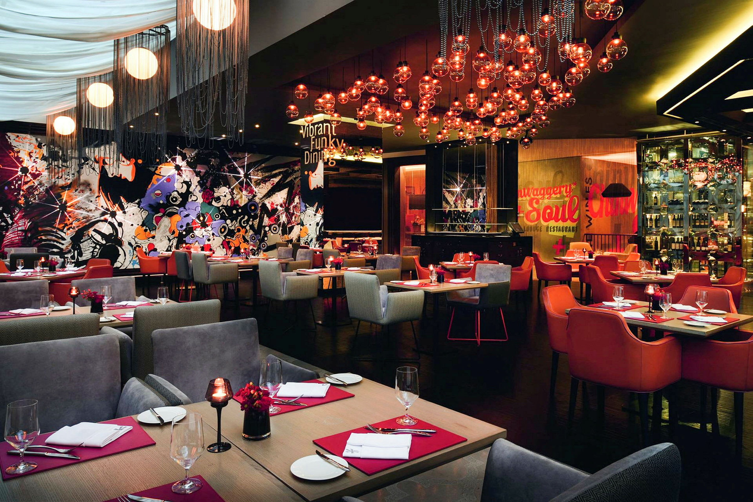 Restaurant med fargerik og moderne innredning, bord med røde servietter og glass samt lamper som henger fra taket. (AI-generert tekst)