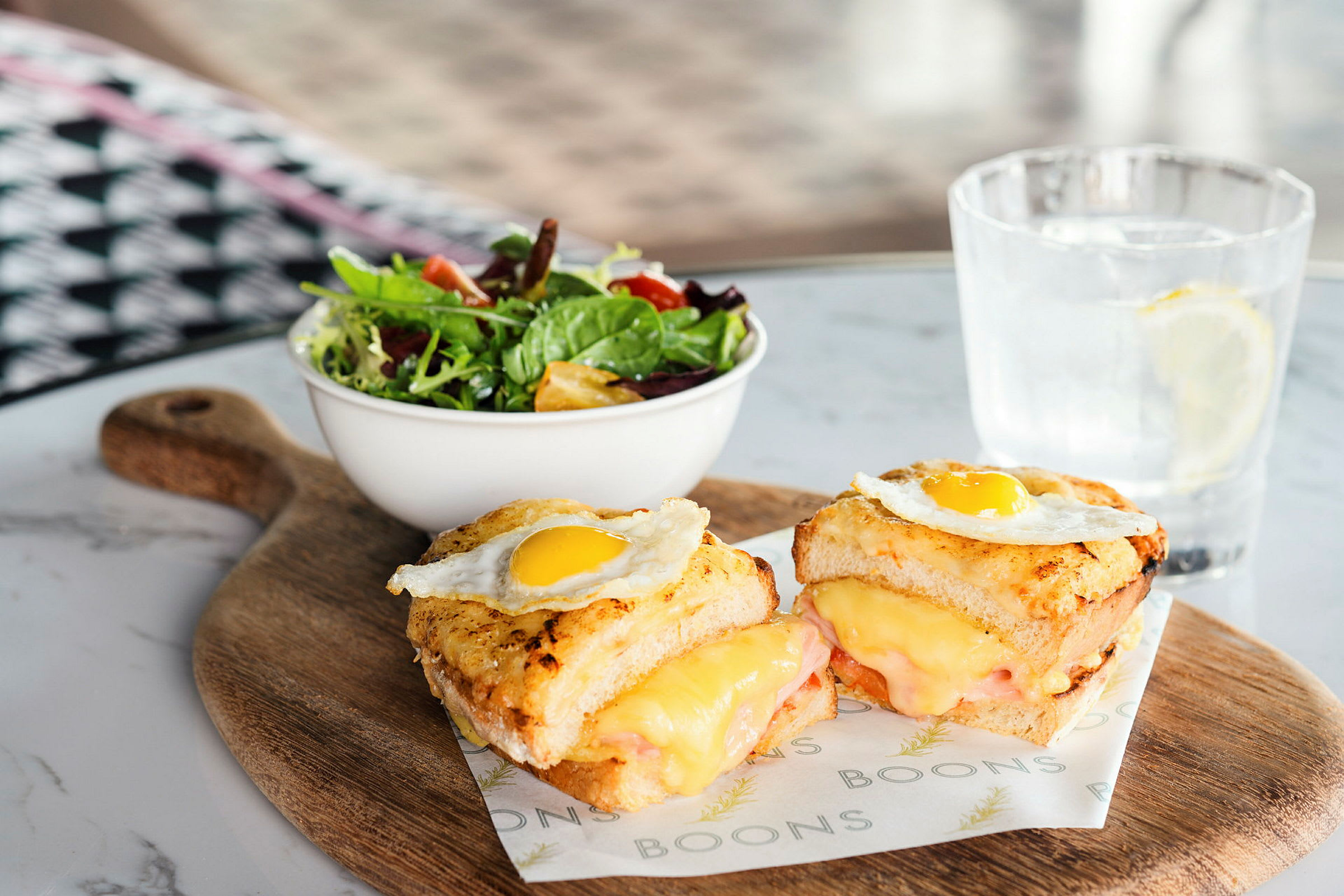 Et trefat med to toast fylt med smeltet ost og toppet med stekt egg, en bolle med salat og et glass vann med sitron. (AI-generert tekst)