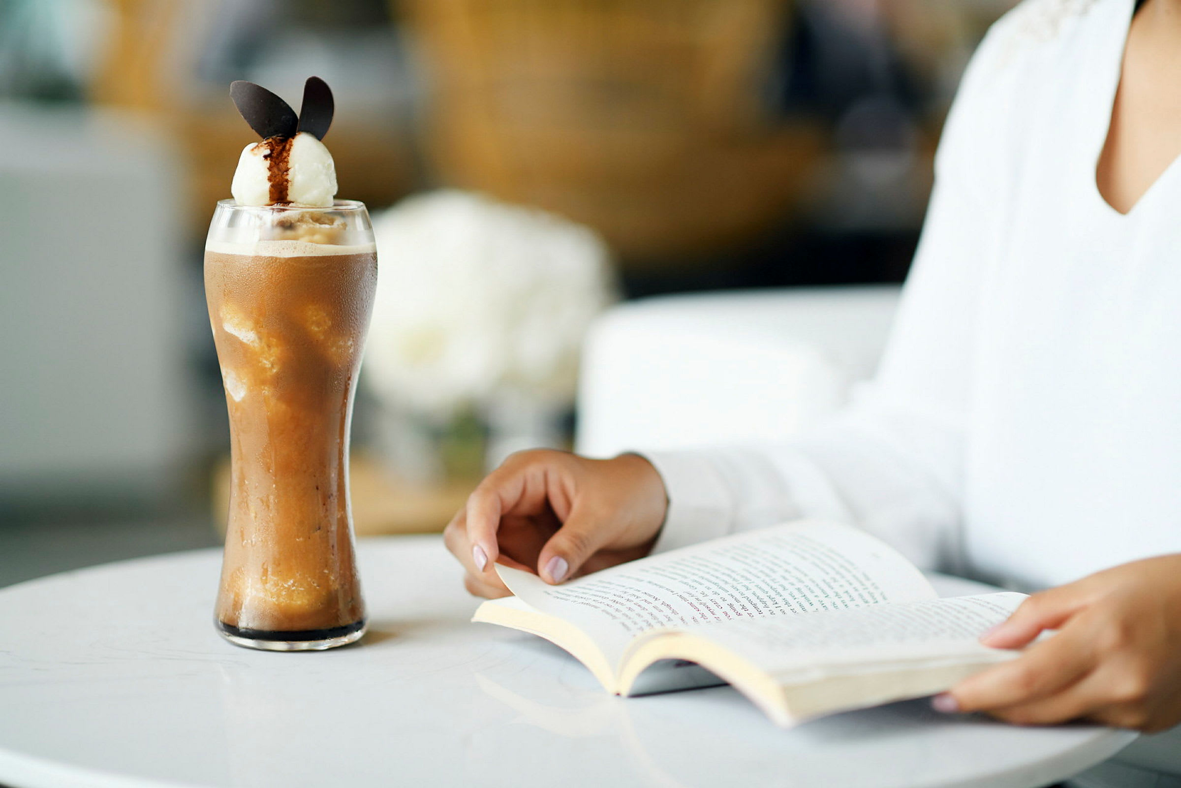Et glass med kald kaffedrikk og en person som leser en bok ved et bord. (AI-generert tekst)