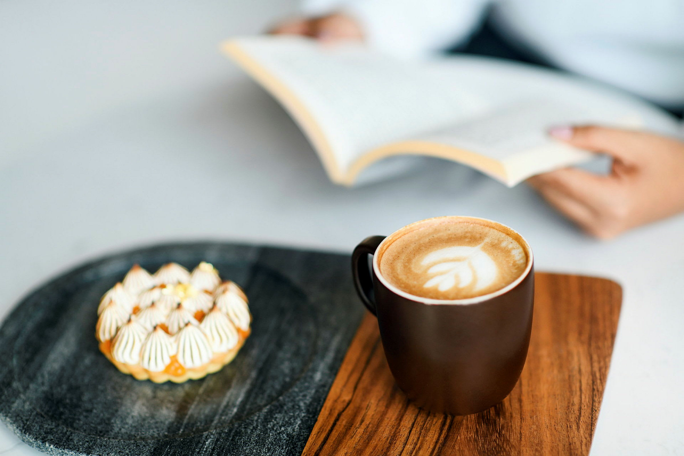 En kopp kaffe og en liten kake ligger på et trebord. En person holder en åpen bok i bakgrunnen. (AI-generert tekst)