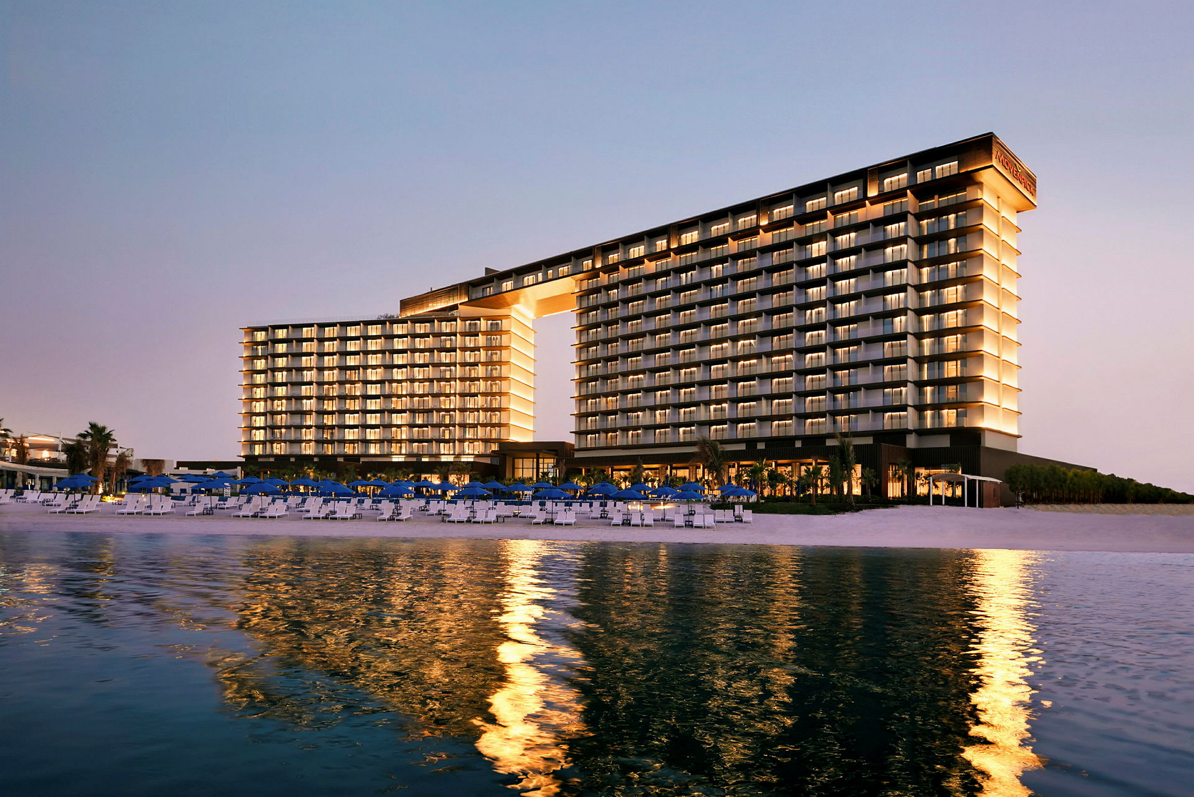 Bygningen er et moderne hotellkompleks ved en strand, opplyst og speiles i vannet. (AI-generert tekst)