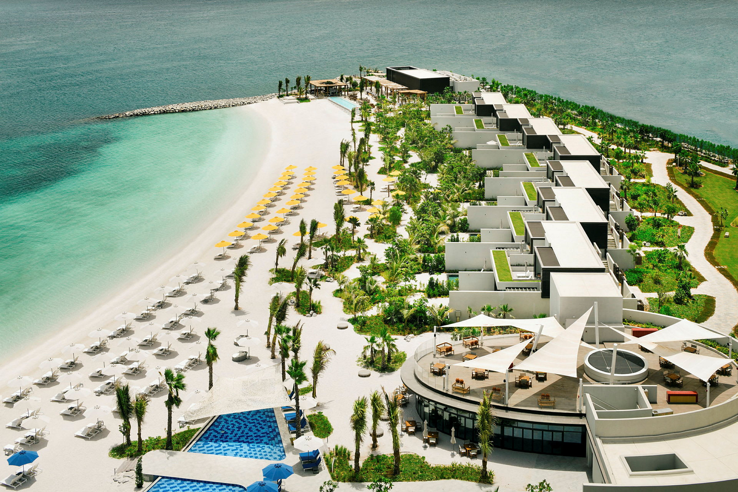 Luftfoto av strand med solsenger og parasoller foran hotellområde med grøntområde. (AI-generert tekst)