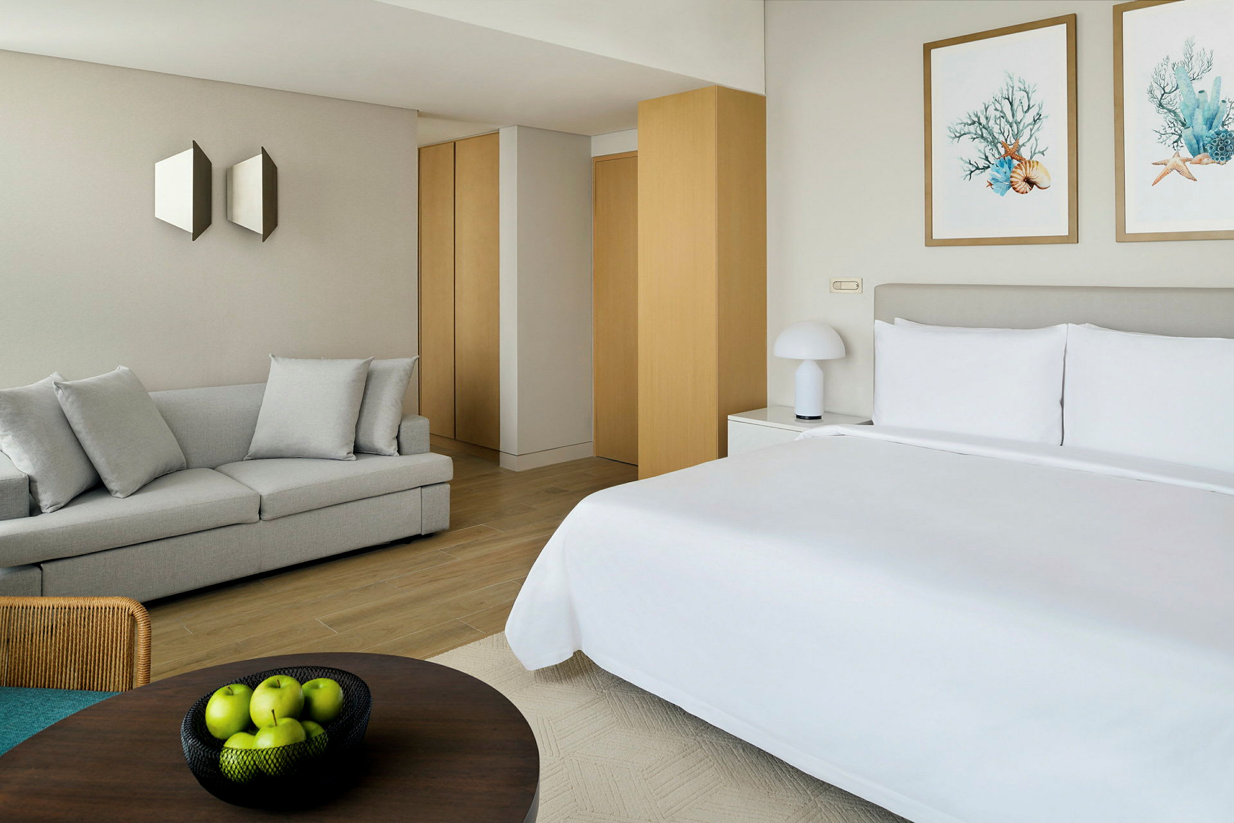 Et moderne hotellrom med en oppredd dobbeltseng. Rommet har også en lys sofa, veggdekorasjoner og en skål med grønne epler på et bord. (AI-generert tekst)