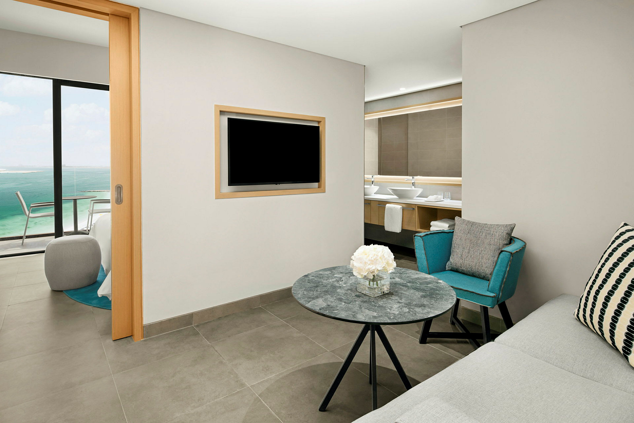 Et hotellrom med sittegruppe, et bord med blomster, en sofa, en lenestol og en TV montert på veggen. I bakgrunnen ses et bad og gjennom en glasskyvedør en utsikt over havet. (AI-generert tekst)