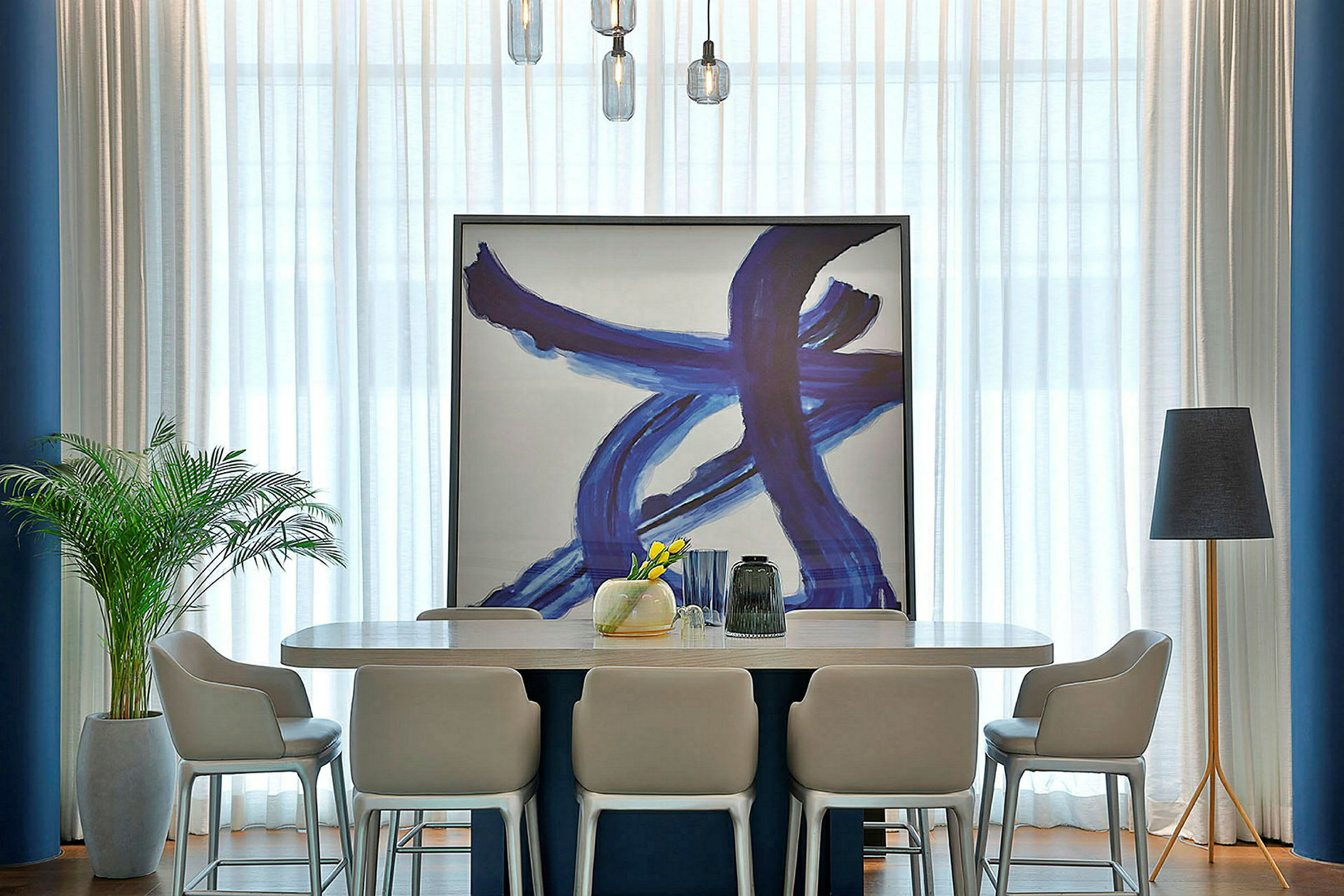 Et interiør med et moderne spisebord omringet av stoler. Et stort kunstverk med blå penselstrøk står lent mot veggen. En gulvlampe og en innendørsplante er plassert på hver side av bordet. (AI-generert tekst)