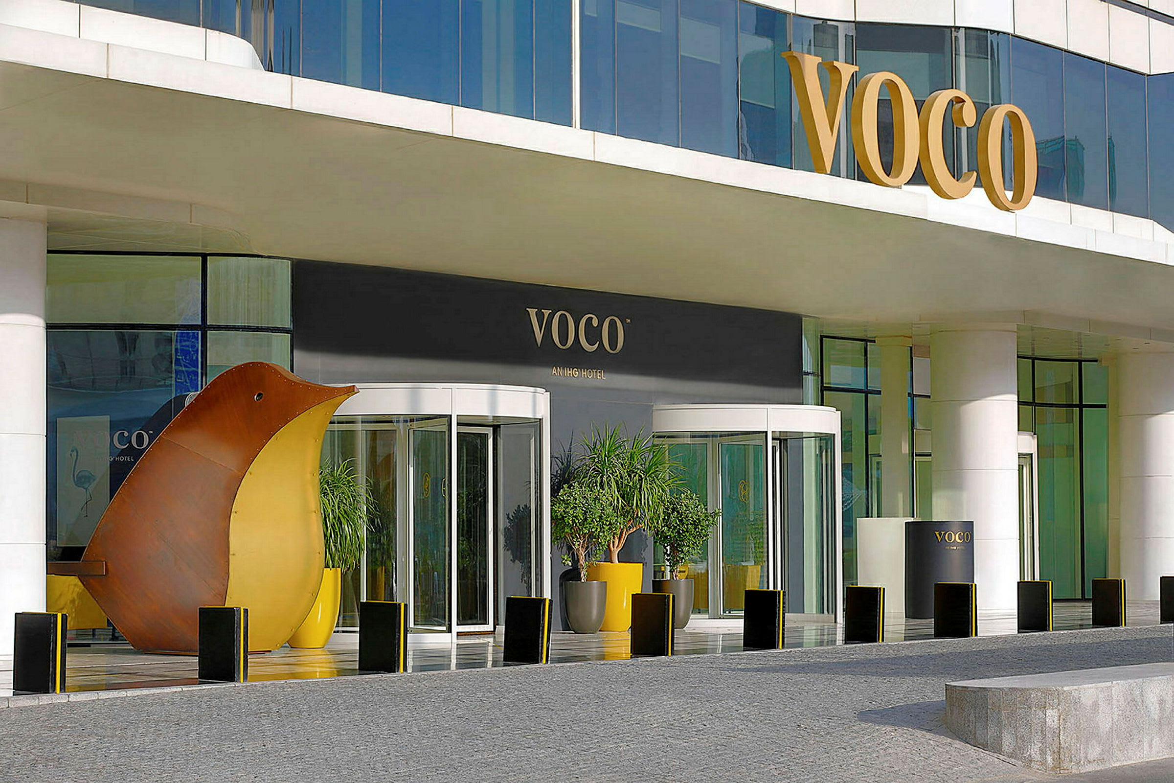 Inngang til et hotell med glassdører og skiltet 'VOCO', samt dekorasjoner og planter. (AI-generert tekst)