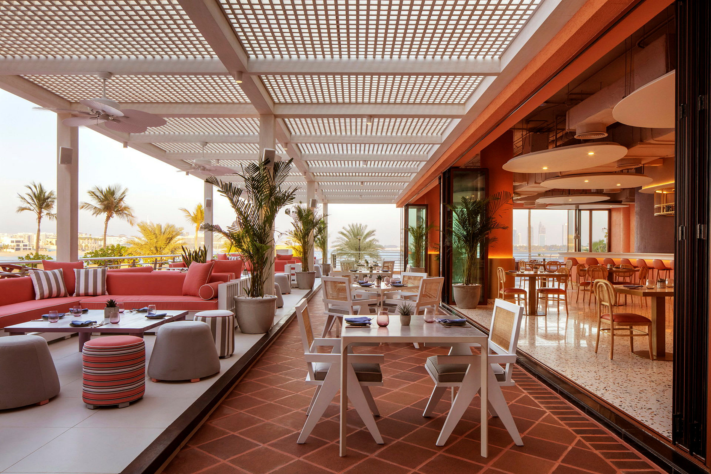 En åpen terrasse på en restaurant med sittegrupper og bord, omgitt av palmer og en luftig atmosfære. (AI-generert tekst)