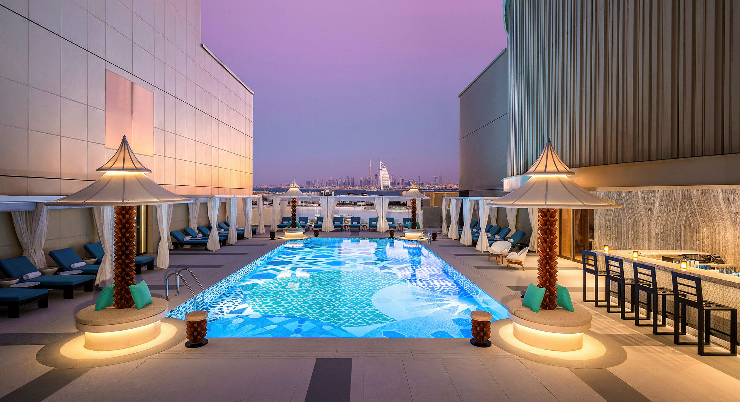 En takterrasse med et dekorativt basseng omgitt av solsenger og små parasoller. I bakgrunnen sees Burj Al Arab og Dubais skyline under en lilla kveldshimmel. (AI-generert tekst)