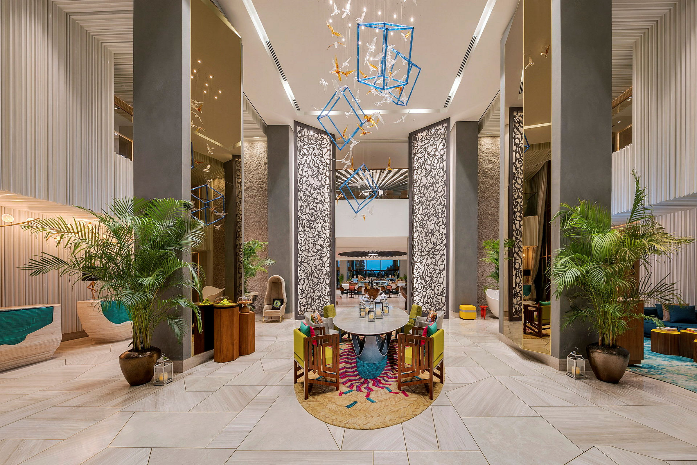 Et moderne og elegant hotellobby med høyt tak, dekorative paneler, hengende moderne installasjoner og planter. Bord og stoler står på et fargerikt teppeområde. (AI-generert tekst)