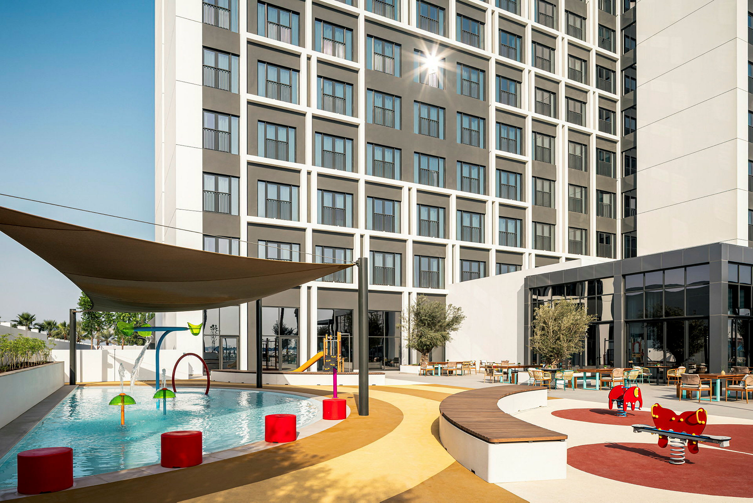 Utendørsområde med barnebasseng, lekeplass og sitteplasser foran et hotell. (AI-generert tekst)