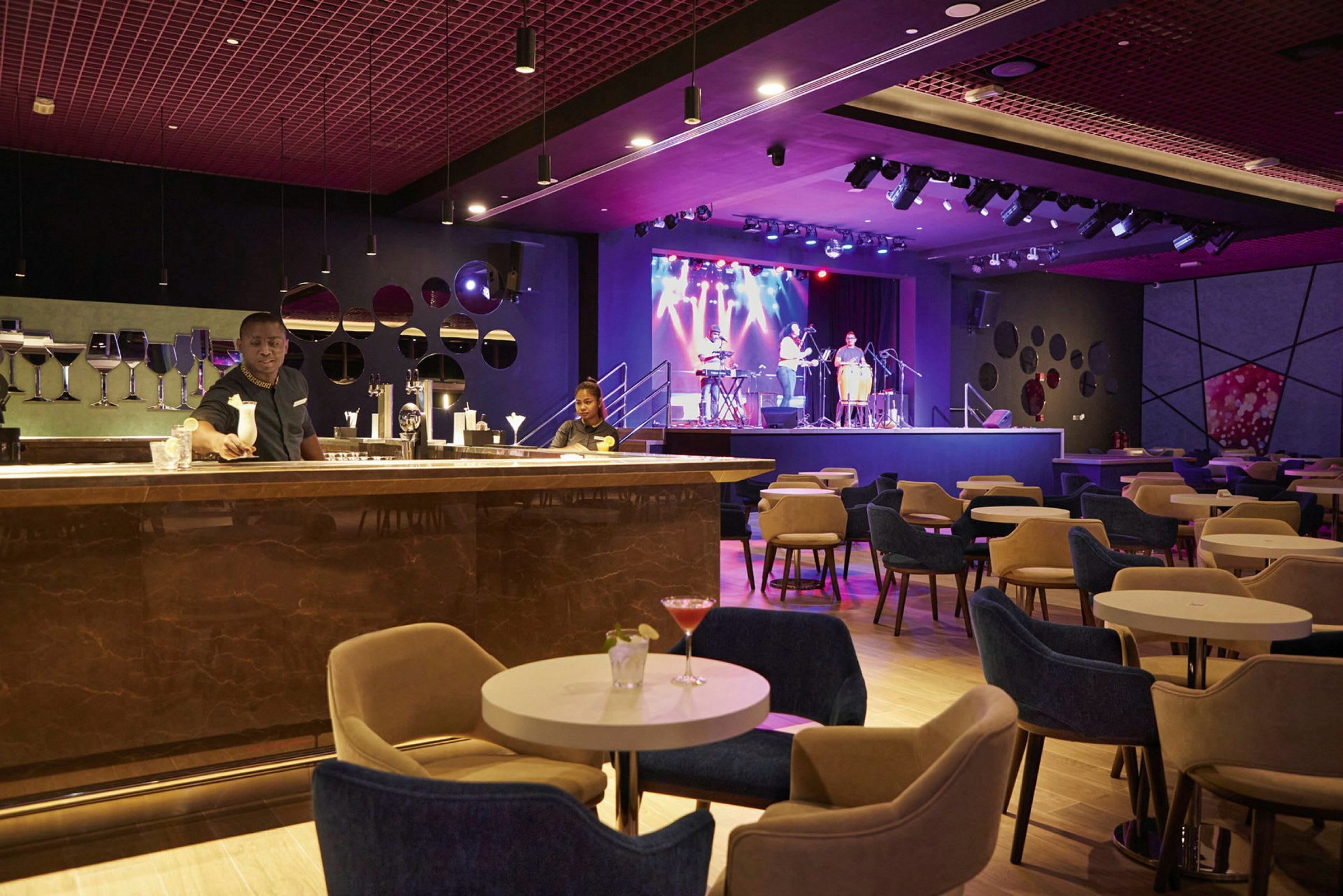 En elegant lounge med sitteplasser rundt små bord, en bardisk i forgrunnen og en scene med musikere i bakgrunnen. (AI-generert tekst)