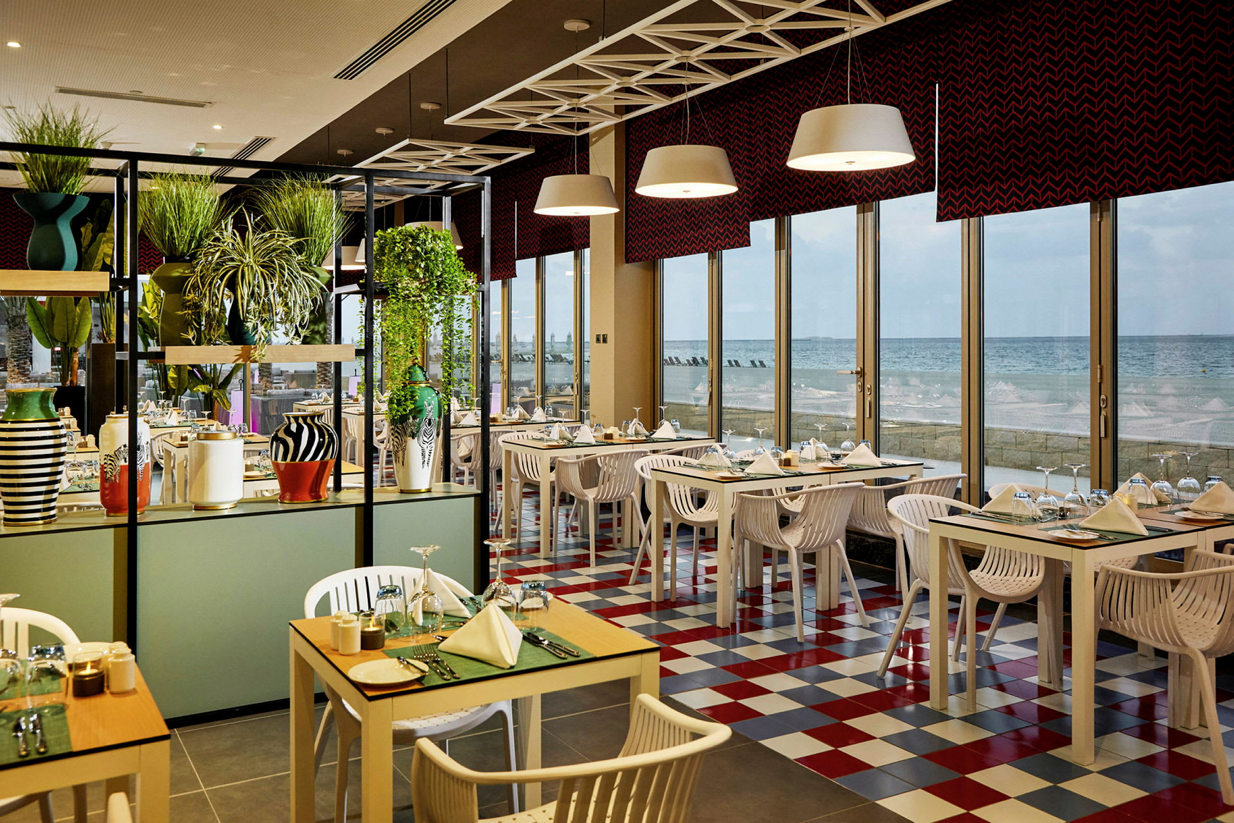 En restaurant med fargerike bord og stoler, store vinduer og utsikt over havet. (AI-generert tekst)