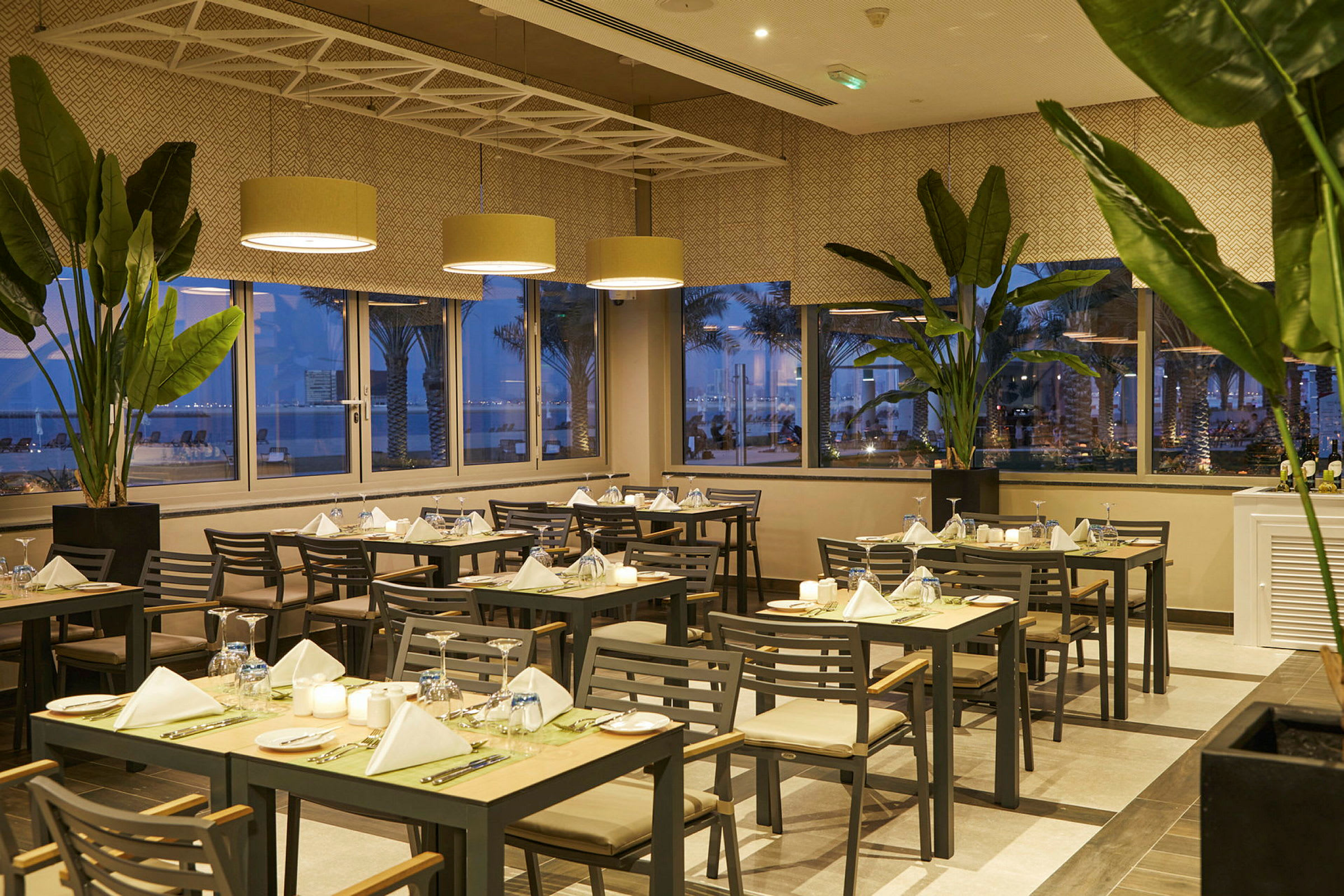 Restaurantmiljø med dekkede bord, grønne planter og panoramavinduer med kveldssutsikt. (AI-generert tekst)