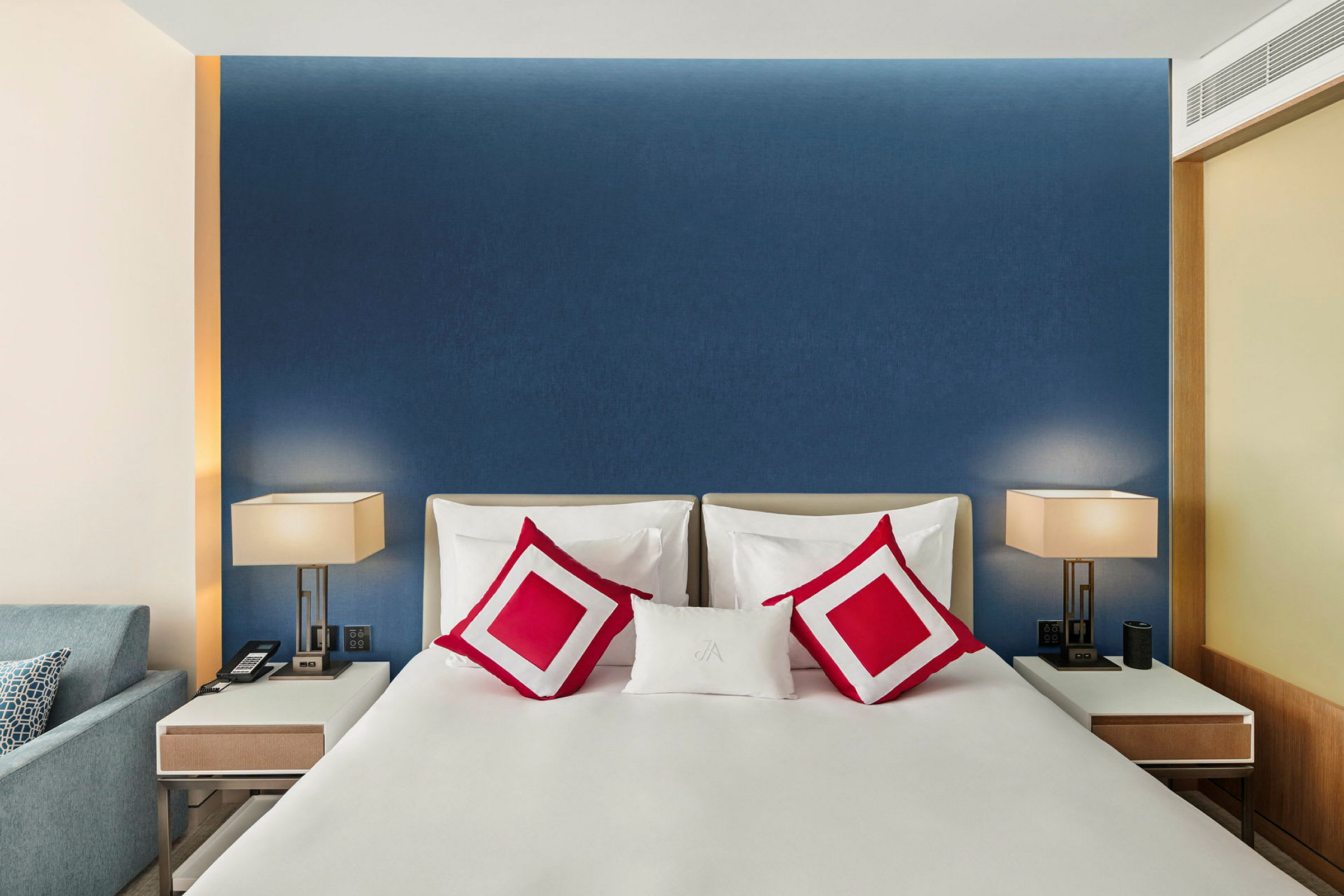 Et moderne, elegant hotellrom med en dobbeltseng som har hvitt sengetøy og røde pynteputer. På hver side av sengen står hvite, stilige nattbord med bordlamper. (AI-generert tekst)
