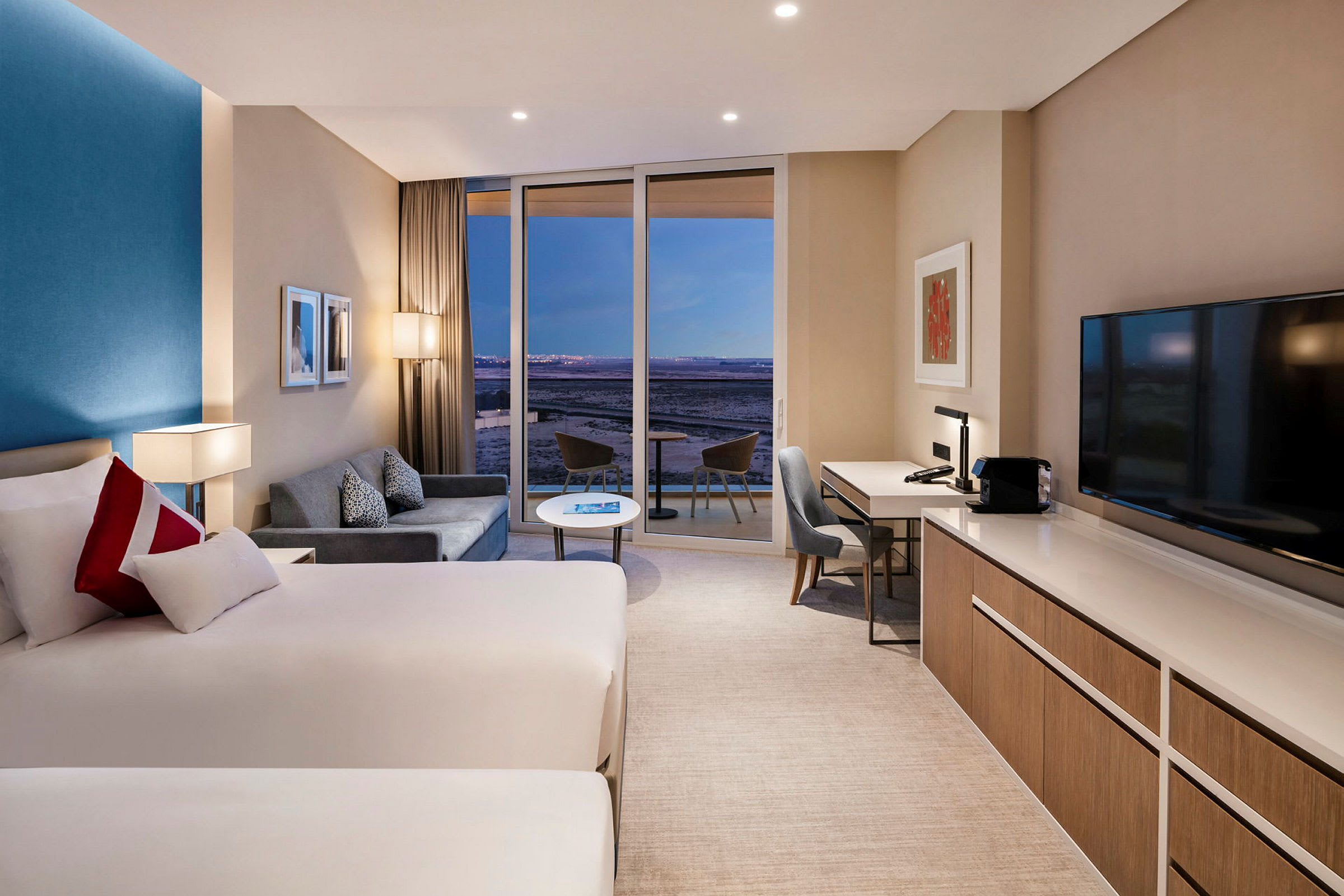 Et moderne hotellrom med to senger, en sofa, skrivebord og en stor glassdør som fører ut til en balkong. (AI-generert tekst)