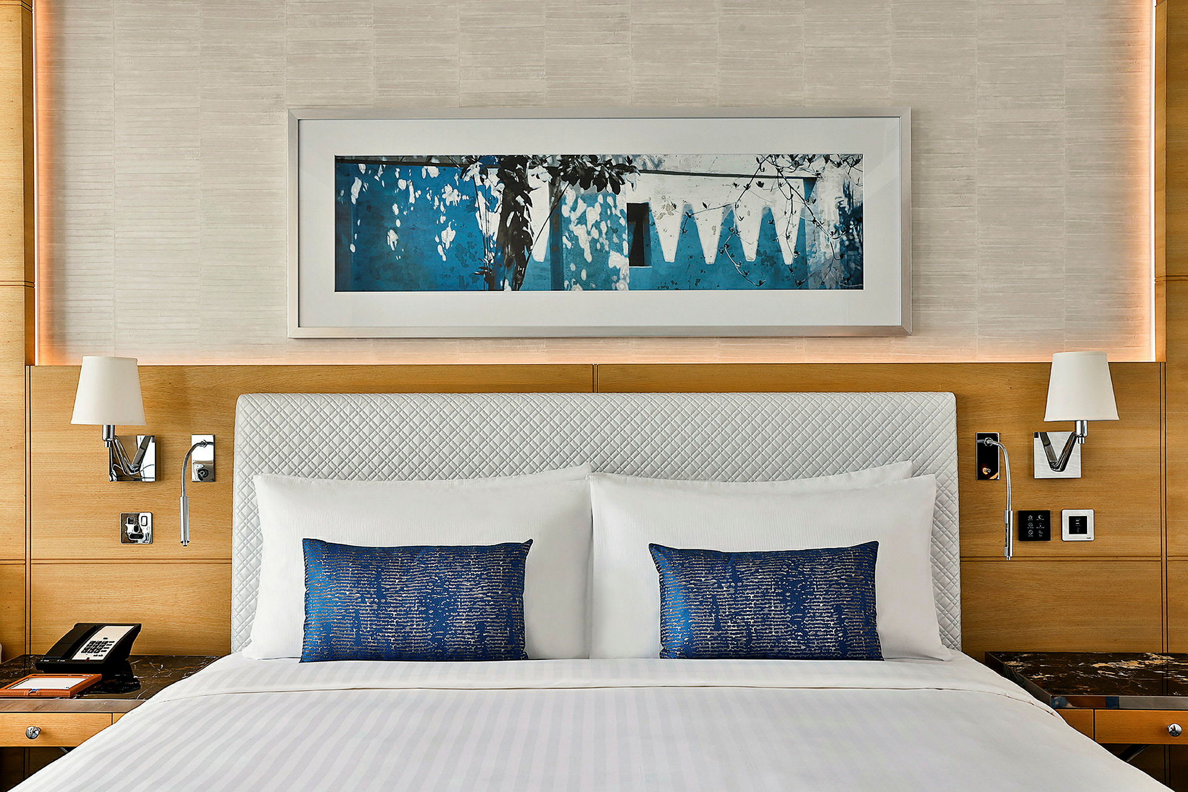 Bilde av en hotellseng med hvitt sengetøy og blå puter, omgitt av en innredning med trepanel og en veggkunst over. (AI-generert tekst)
