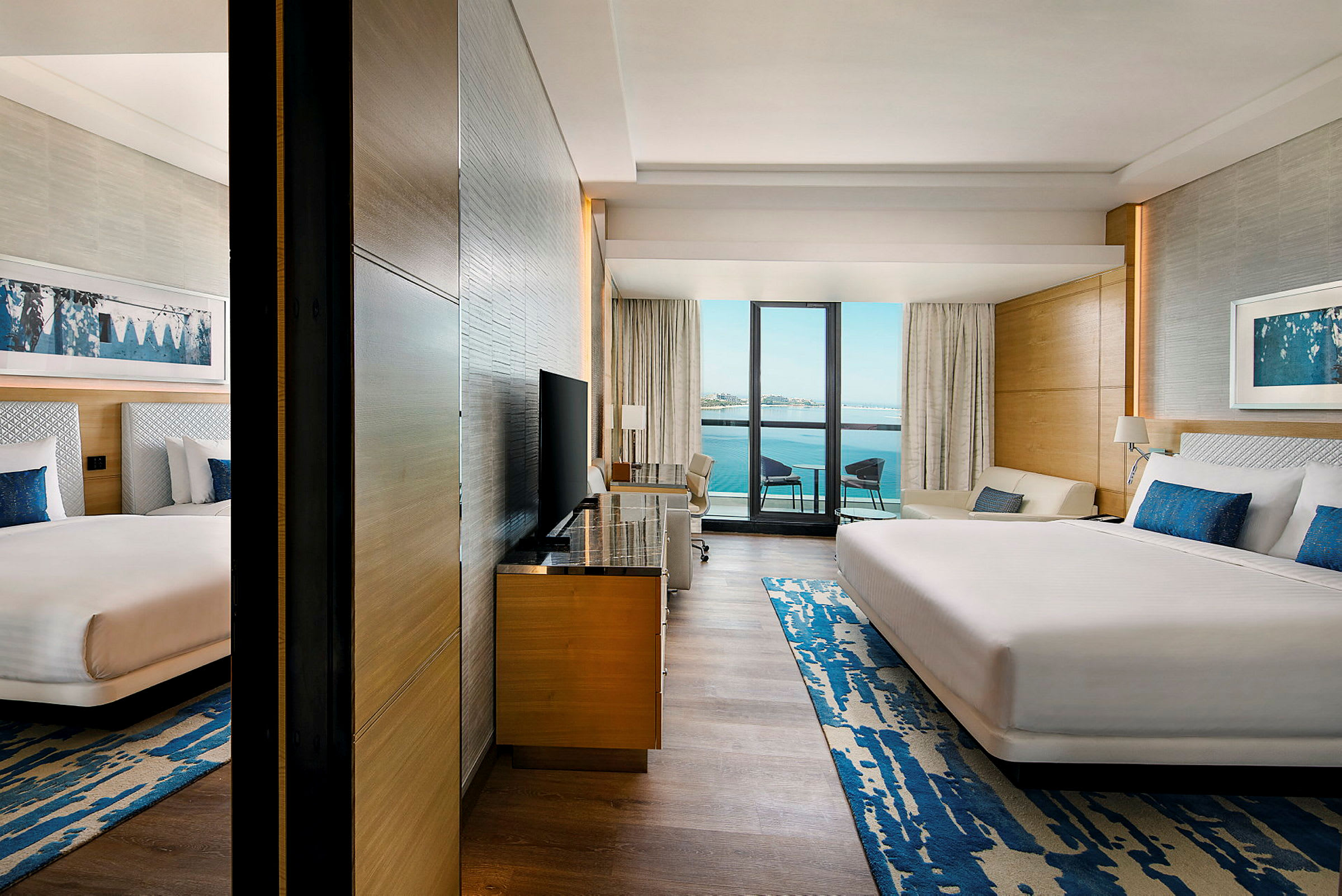 En hotellsuite med to senger, sitteplasser og en balkong med utsikt over en vannflate. (AI-generert tekst)