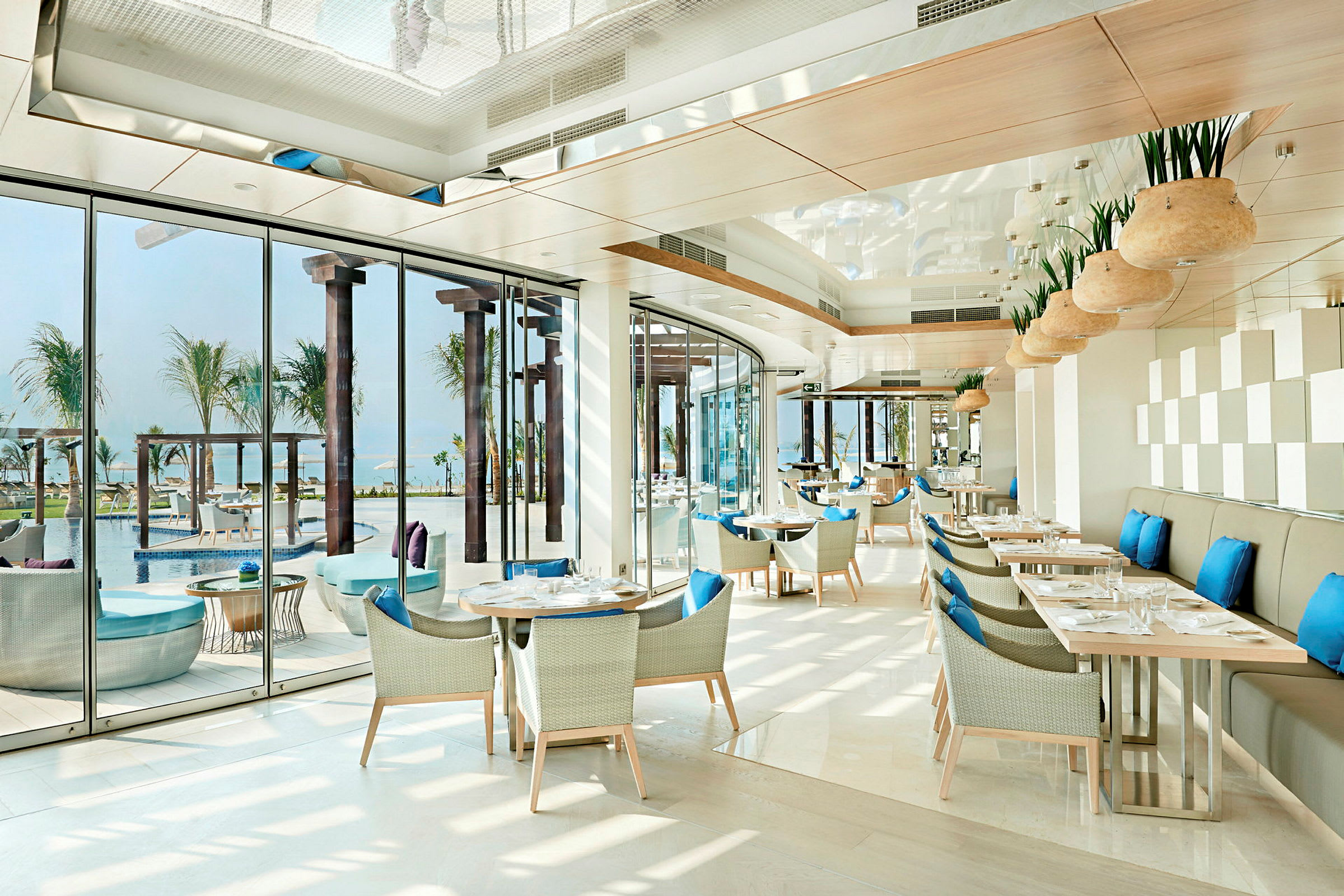 En lys og moderne restaurant med blå puter på stolene og utsikt over et basseng og palmer. (AI-generert tekst)