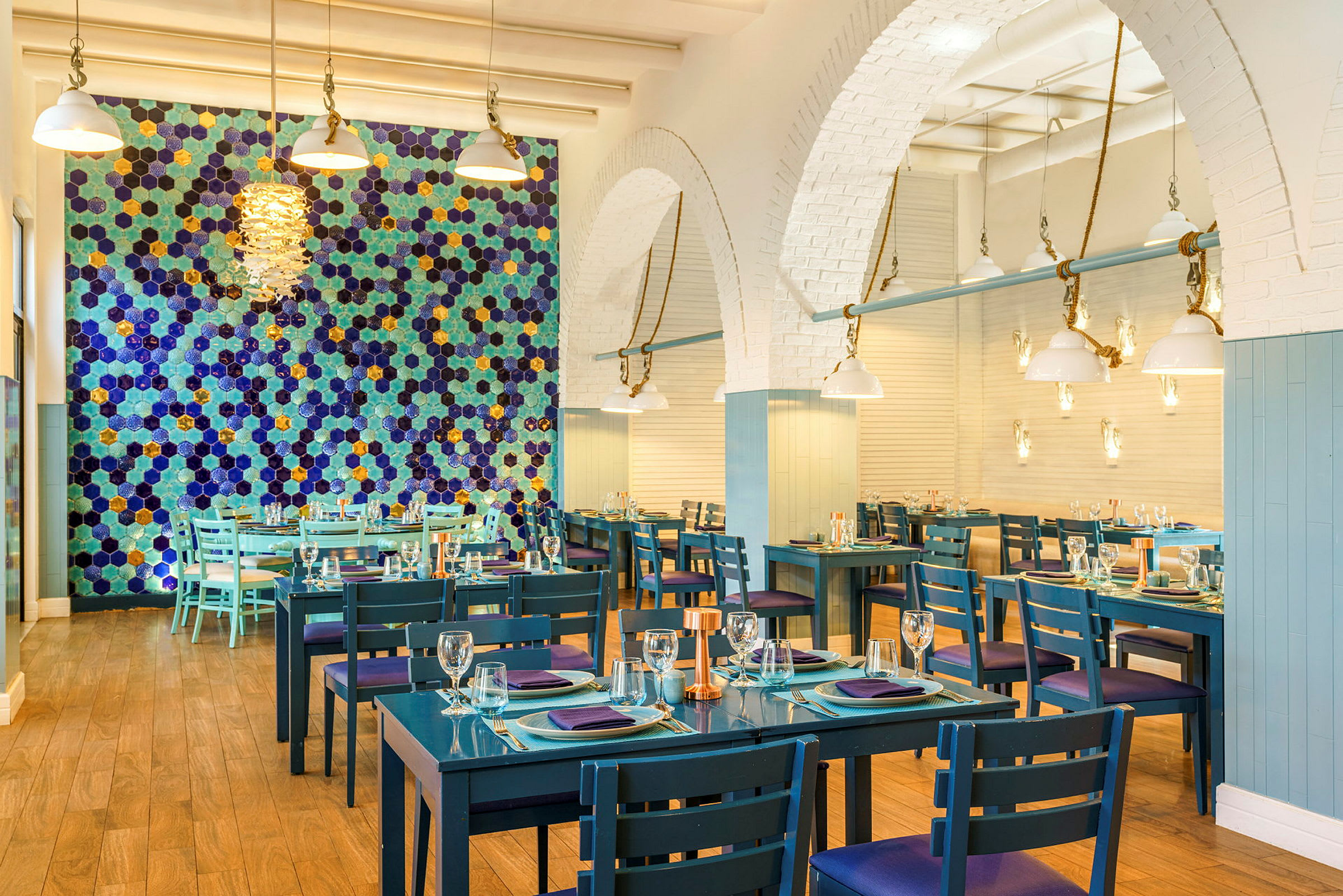 Bilde av en restaurant med blå bord og stoler, en vegg med fargerike heksagonale mønstre, og moderne taklamper. (AI-generert tekst)