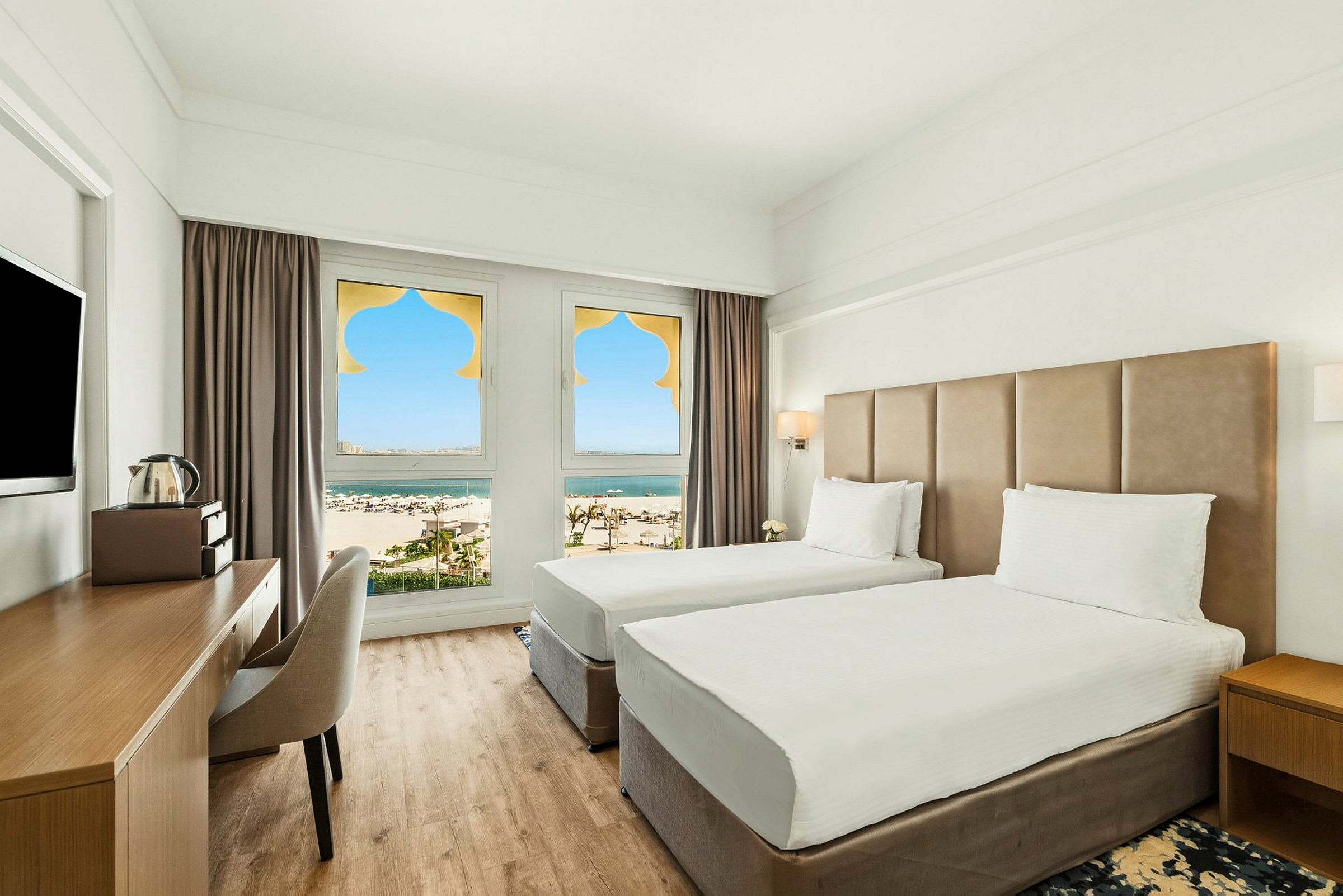 Et hotellrom med to enkeltsenger, en skrivebordplass og vinduer med utsikt over en sandstrand og hav. (AI-generert tekst)