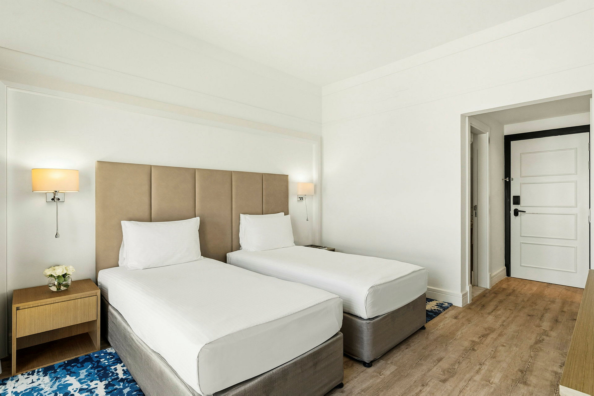 Et hotellrom med to enkeltsenger, et nattbord og en dør. (AI-generert tekst)