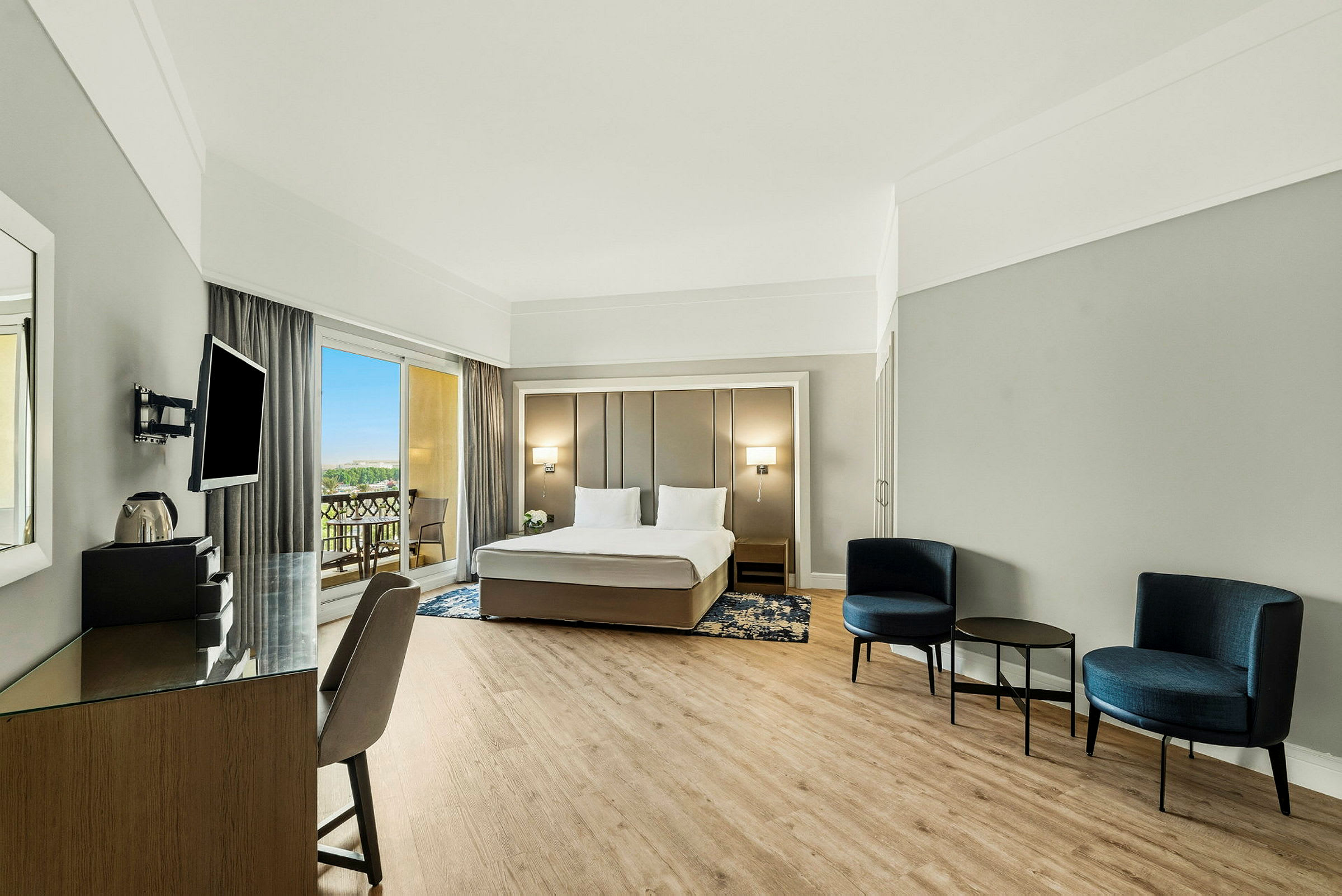 Et moderne hotellrom med en dobbeltseng, en sittegruppe med stoler og bord, samt en arbeidsplass. Utgang til balkong synlig gjennom store vinduer. (AI-generert tekst)